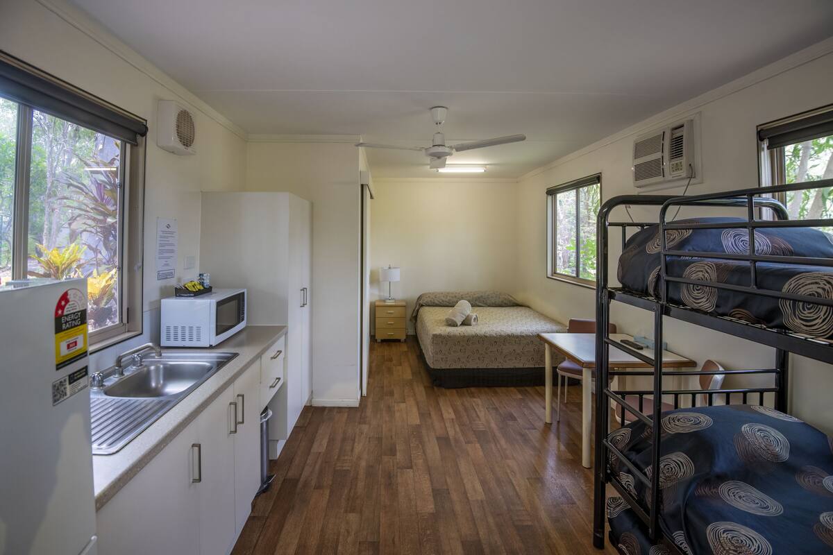 Cocina compacta con microondas, pileta y alacenas. Cuenta con una cama plegable con ropa de cama estampada y una mesita con una silla.