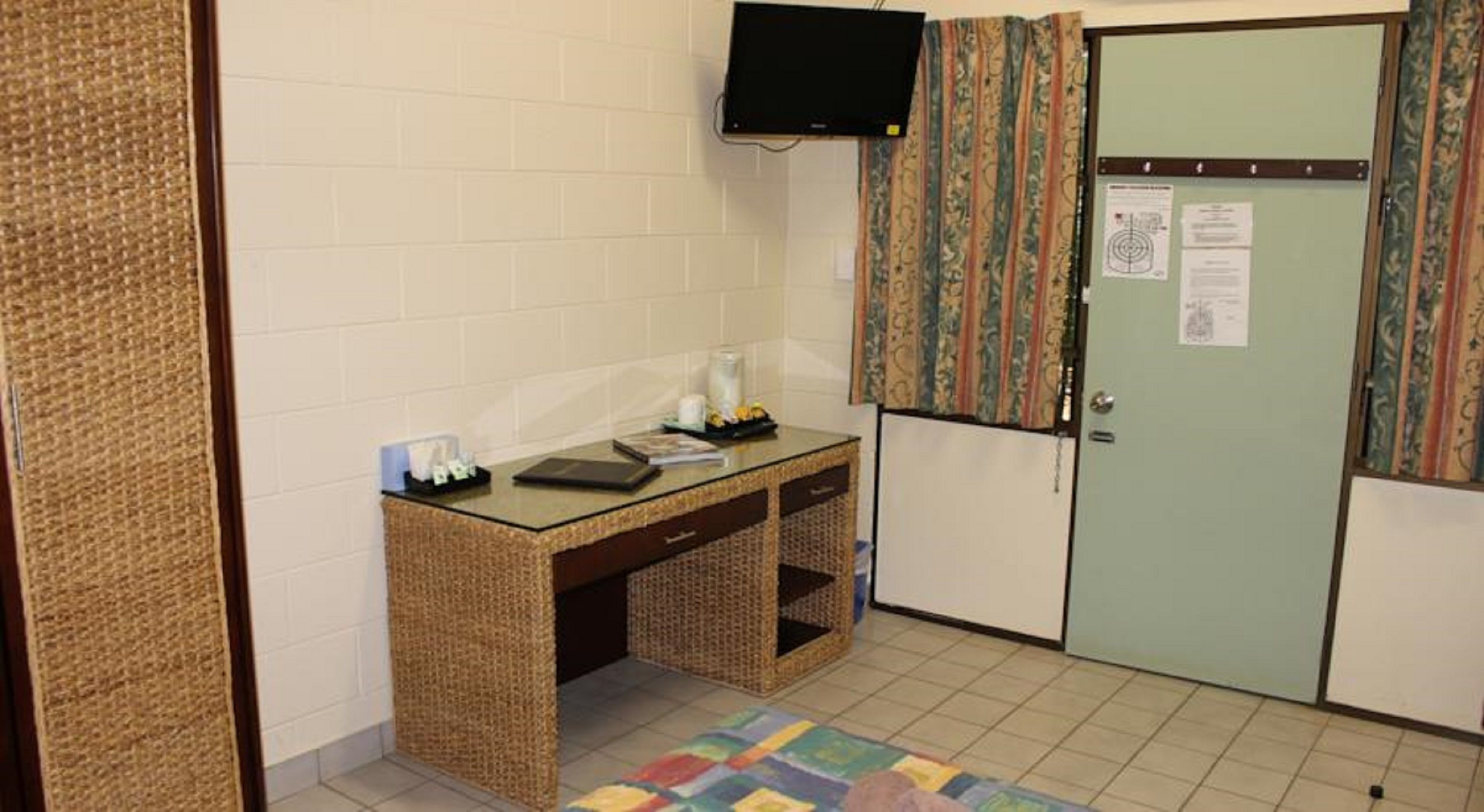 A small room with a desk, a television mounted on the wall, a door with a notice, and a patterned rug on the floor.