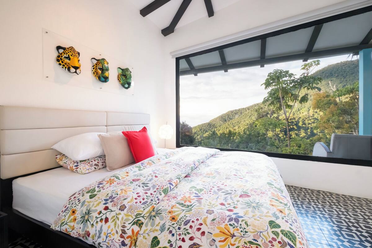 A bedroom with a large bed, floral bedding, a red pillow, and a view of a mountainous landscape through a large window.