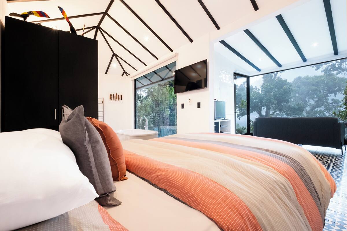 A modern bedroom with a large bed, a black wardrobe, and a glass wall offering a view of the outdoors.