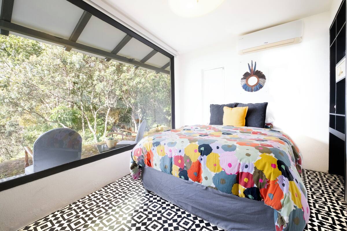 A bedroom with a large window, a bed with a colorful floral bedspread, a black and white patterned floor, and a wall-mounted air conditioning unit.