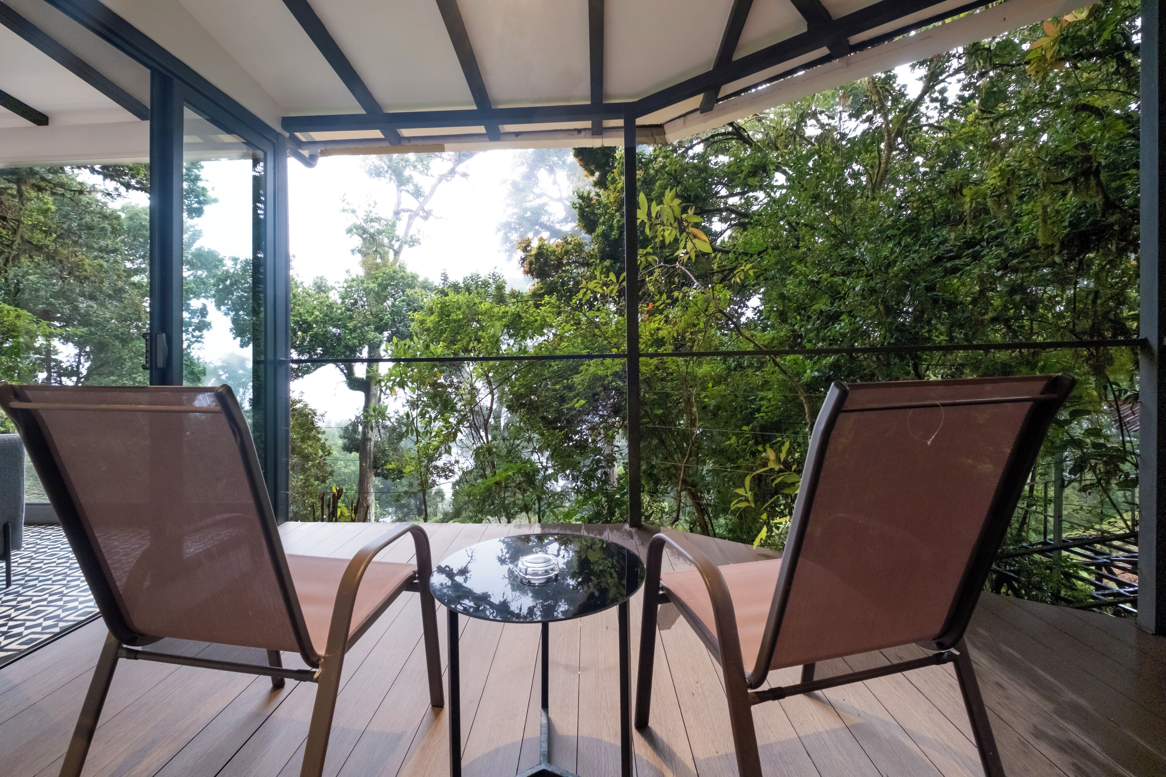 A covered outdoor seating area with two chairs and a table, surrounded by lush greenery and trees.