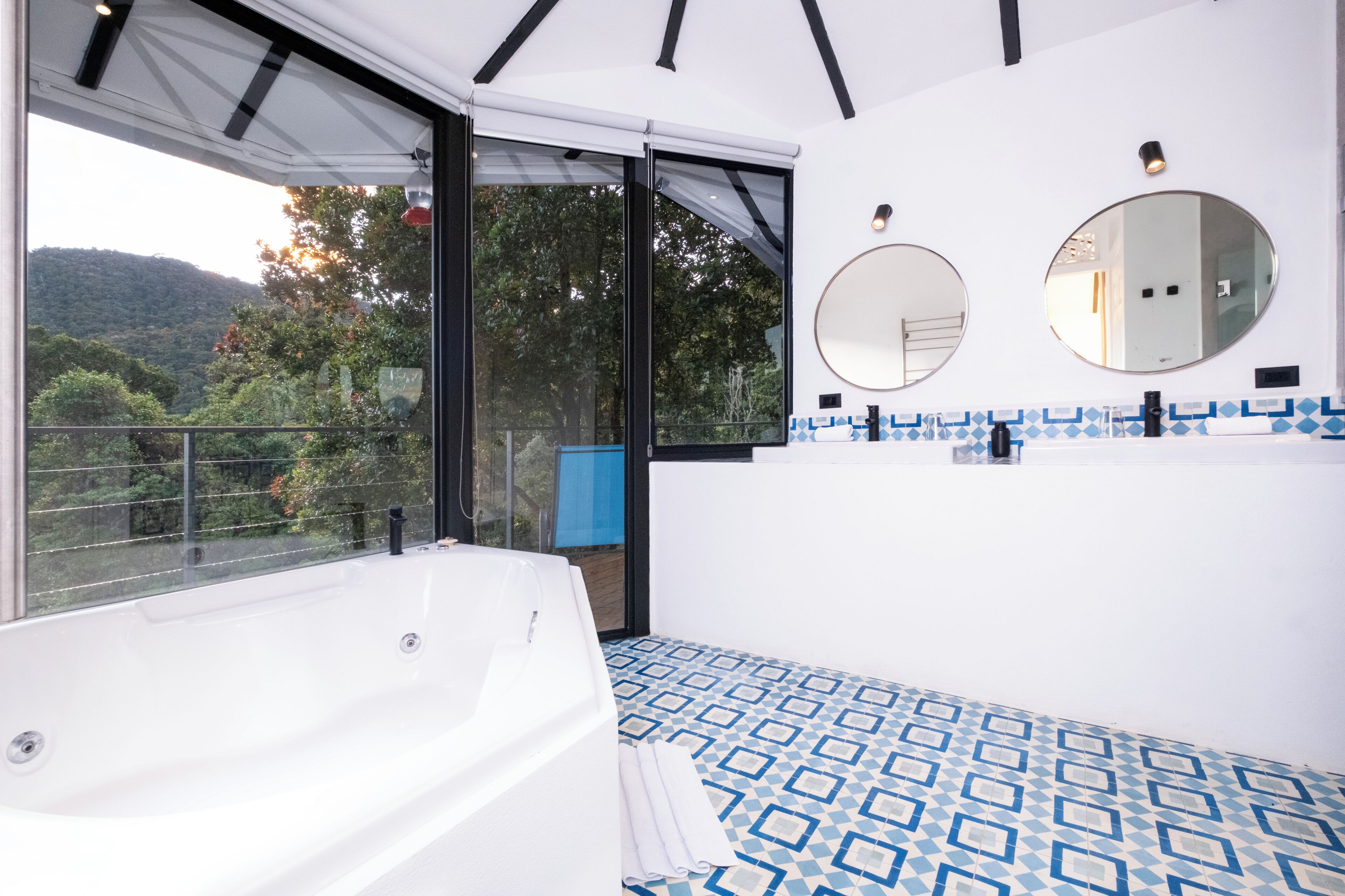 A modern bathroom with a large bathtub, a shower, and a sink. There are two mirrors on the wall, and the room has a large window with a view of trees and hills.