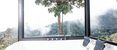 A modern bathroom with a large bathtub, a window with a view of a forested mountain, and a black frame around the window.