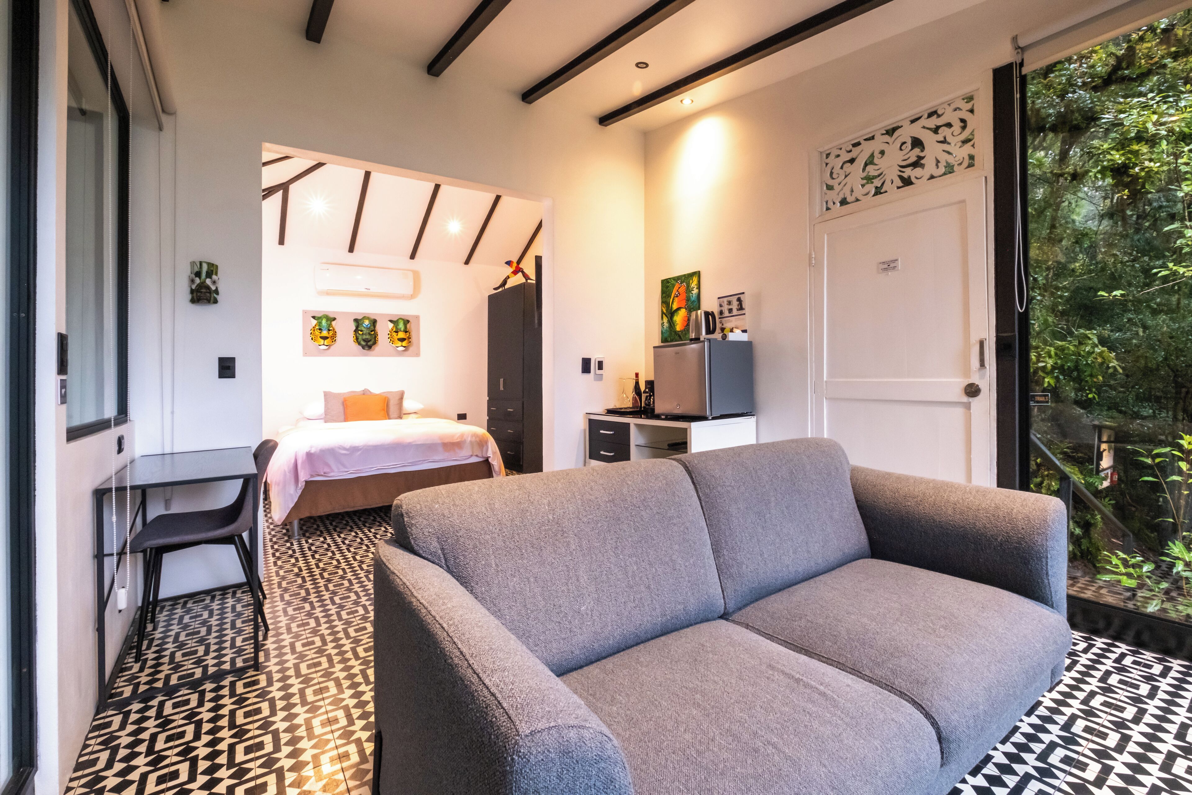 A modern room with a grey sofa, a bed, a desk, and a TV. There is a patterned floor and a door leading to a balcony with greenery.