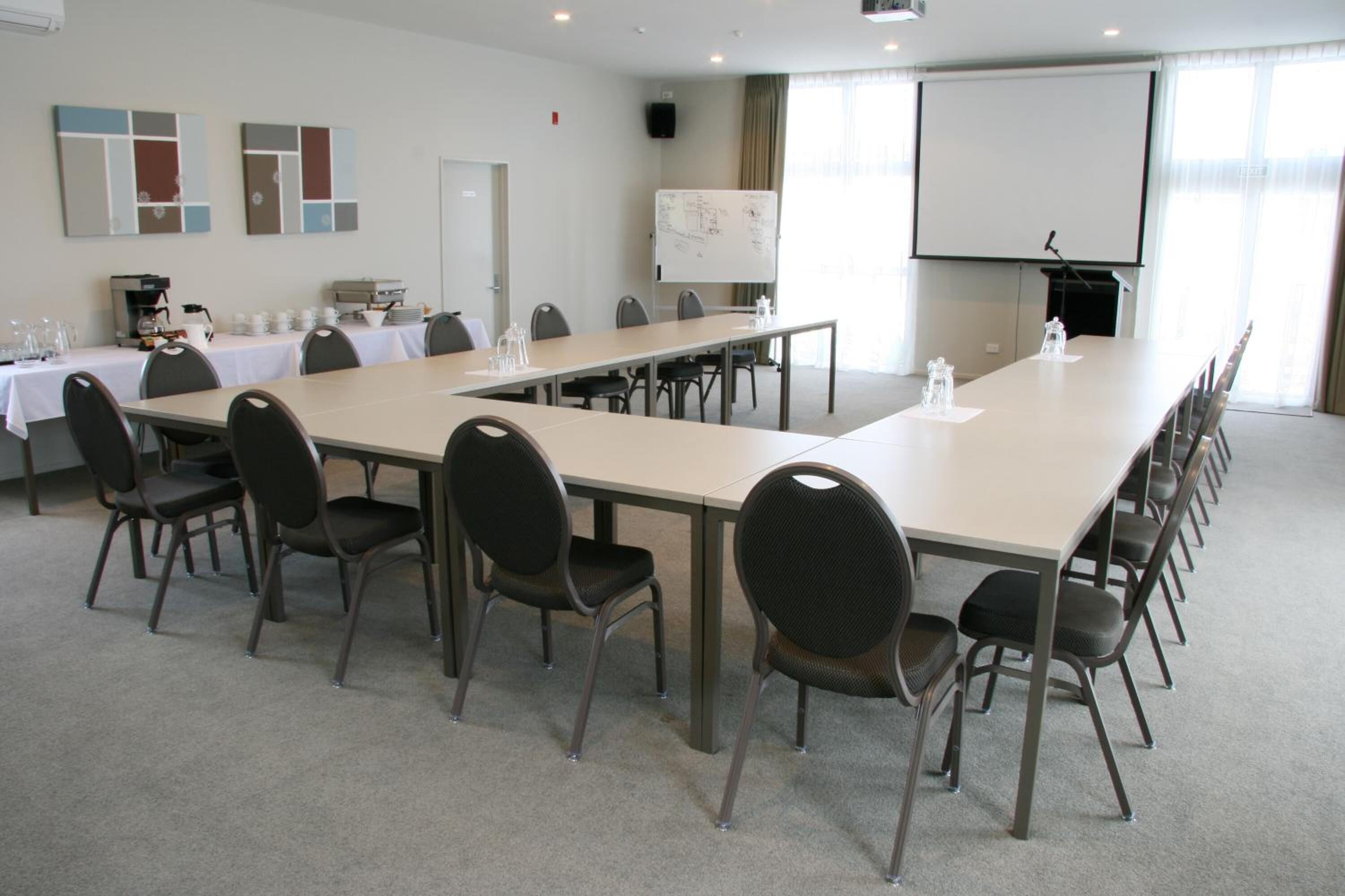 Ein Konferenzraum mit langem Tisch, mehreren Stühlen, einer Whiteboard-Tafel, einer Projektionsleinwand und einer Kaffeestation.