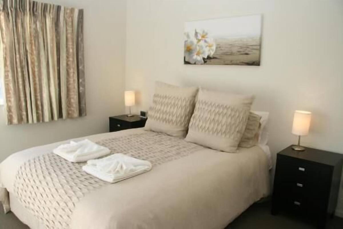 Cama bem arrumada com colcha bege e almofadas, duas mesas de cabeceira com abajures, um quadro na parede e uma janela com cortinas.