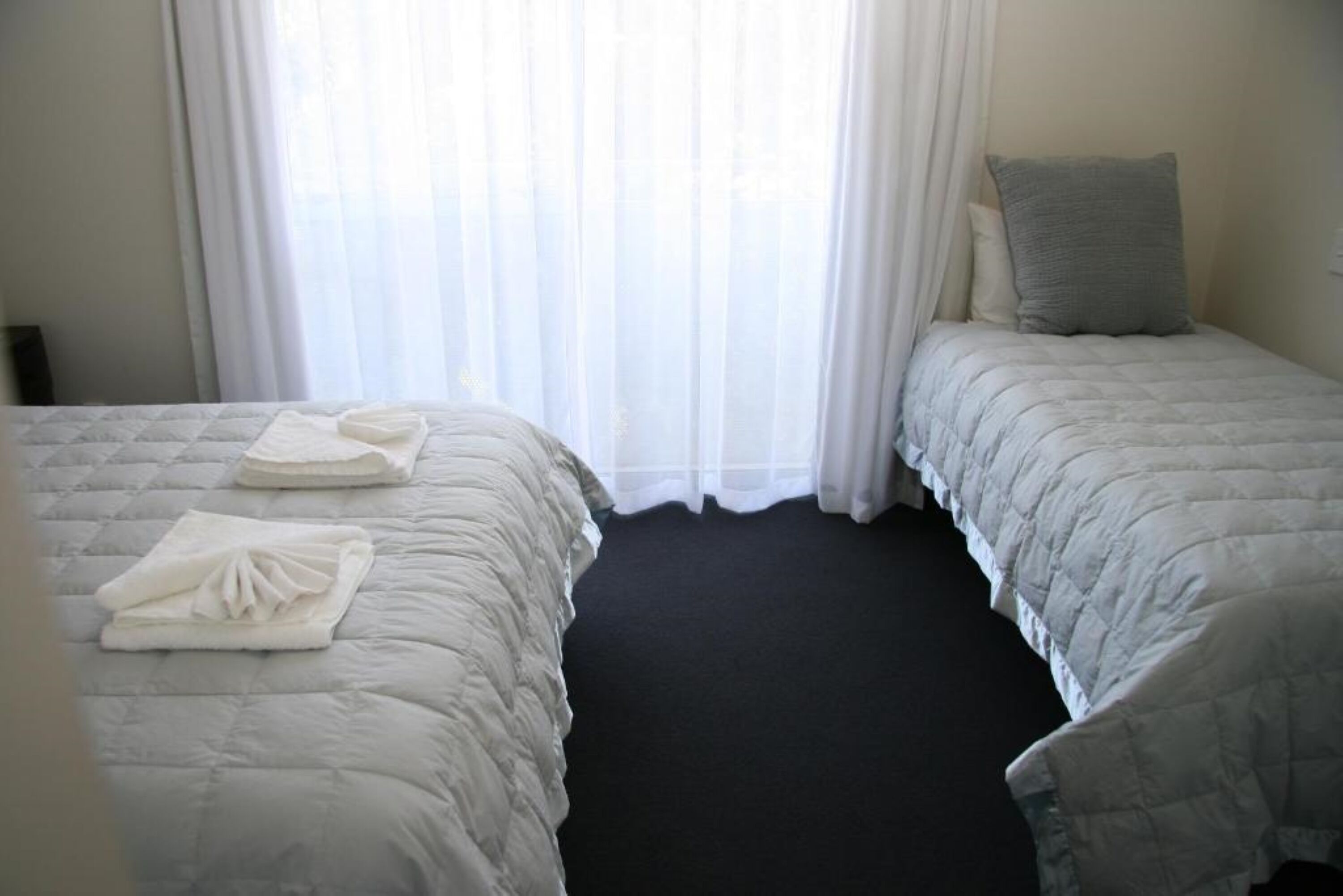 A hotel room with two beds, each with a white duvet and a pillow. There are towels neatly folded on each bed. The room has a dark carpet and a window with sheer curtains.