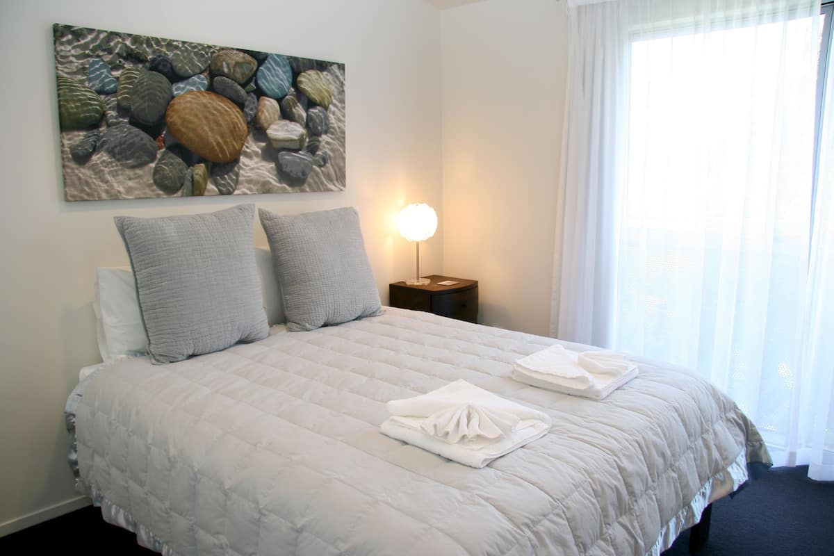 Cama bem arrumada com colcha acolchoada, dois travesseiros e toalhas dobradas em uma mesa de cabeceira com abajur.