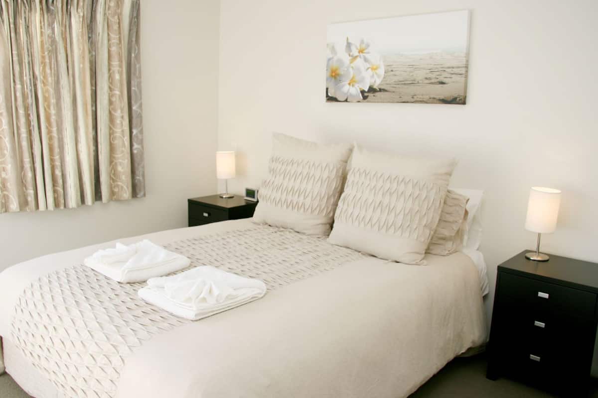 Cama bem arrumada com colcha estampada, duas almofadas e toalhas dobradas. Há duas mesas de cabeceira com abajures, um quadro de flores na parede e uma cortina à esquerda.