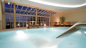 An indoor swimming pool with clear glass ceilings, wooden lounge chairs, and a palm tree. - Hotel Bernstein (Sellin)