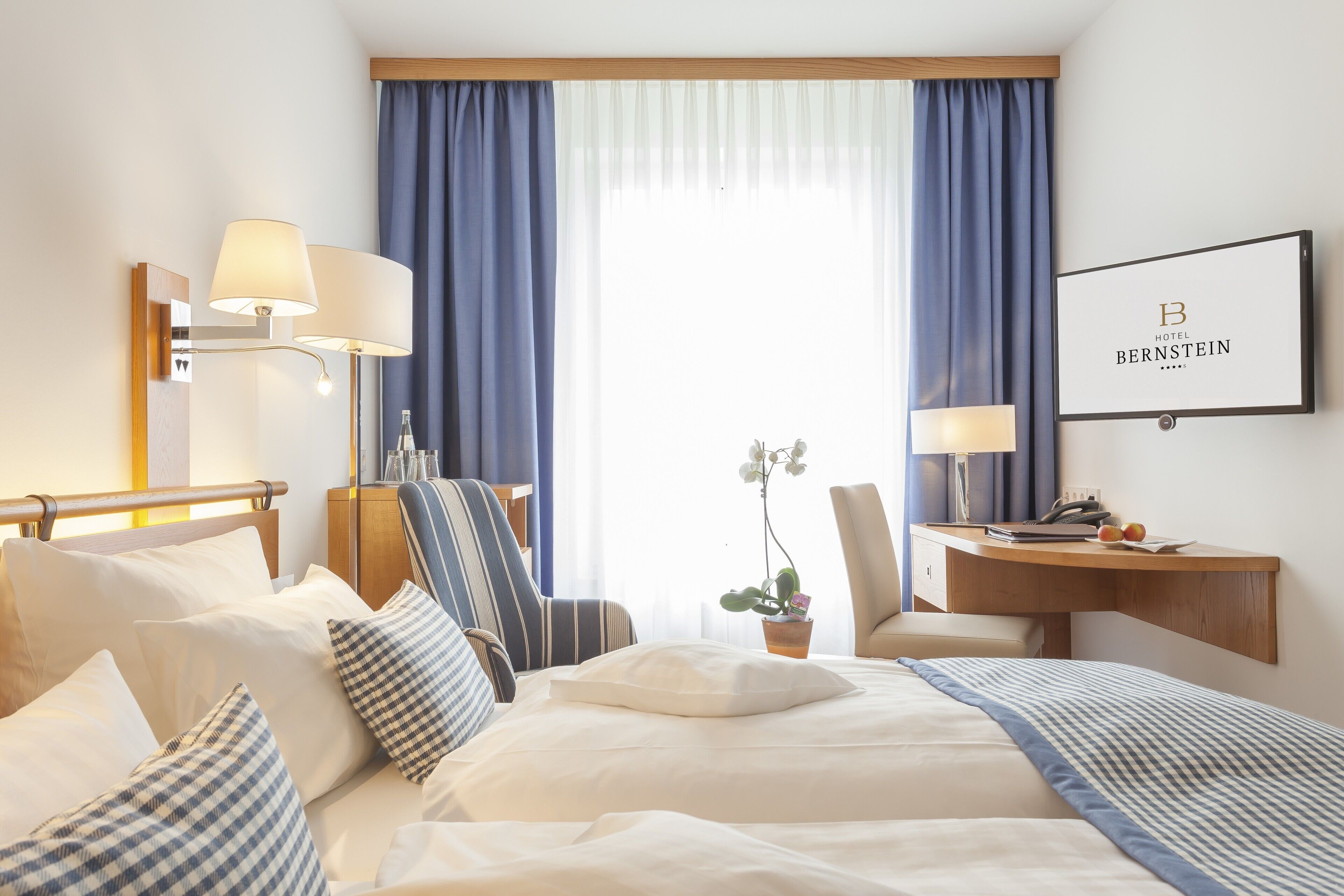A hotel room with a bed, a desk, a chair, a lamp, a potted plant, and a framed sign on the wall.