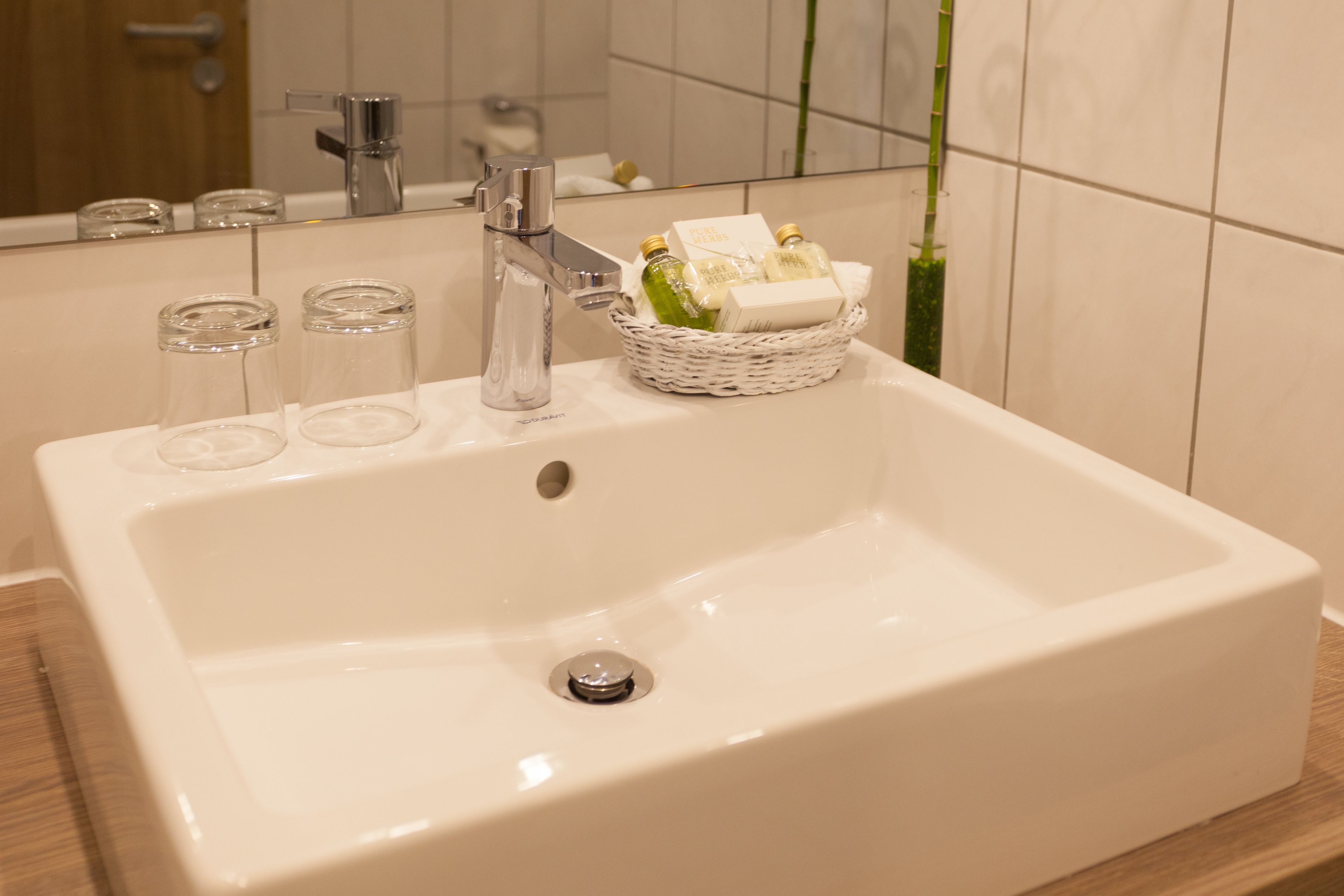 A bathroom with a white sink, a chrome faucet, and a mirror. There are three clear glass bottles and a wicker basket with toiletries.