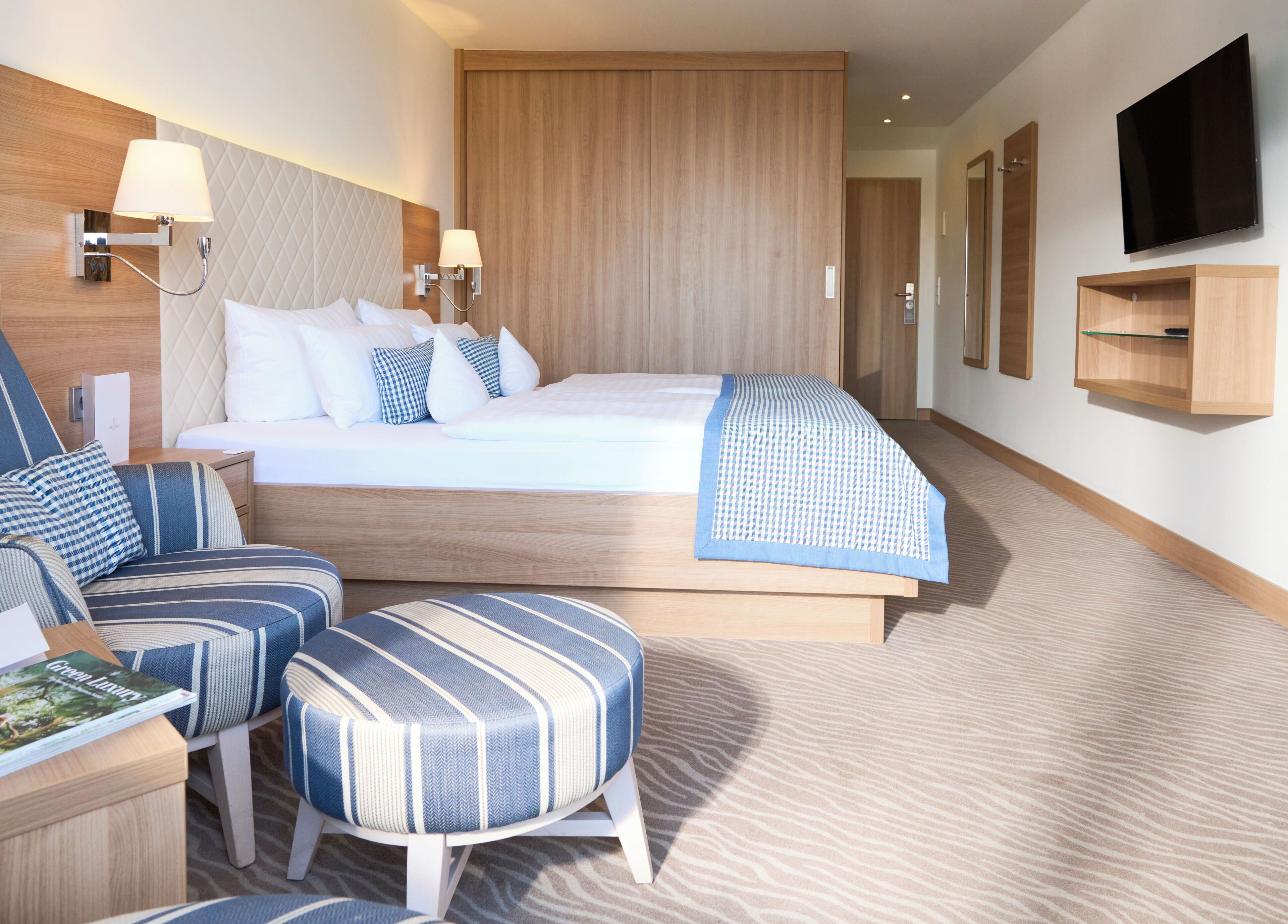 A modern hotel room with a large bed, a seating area with striped chairs, a flat-screen TV mounted on the wall, and a wooden wardrobe.