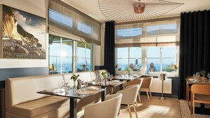A modern dining area with a view of the sea, featuring leather seating, wooden tables, and large windows. - Hotel Bernstein (Sellin)