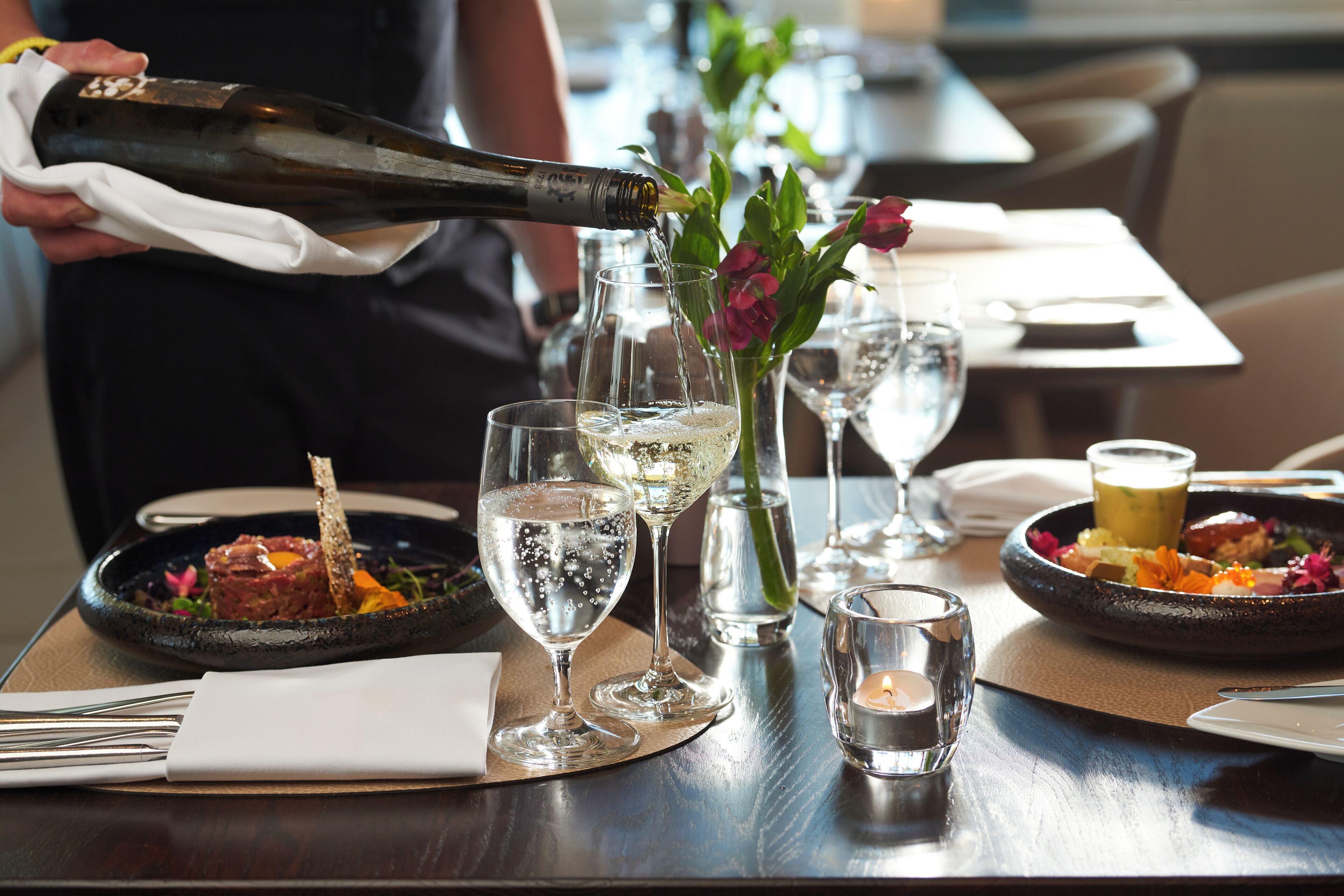 A dining table with a bottle of champagne being poured into a glass, surrounded by plates of food and glasses of water.