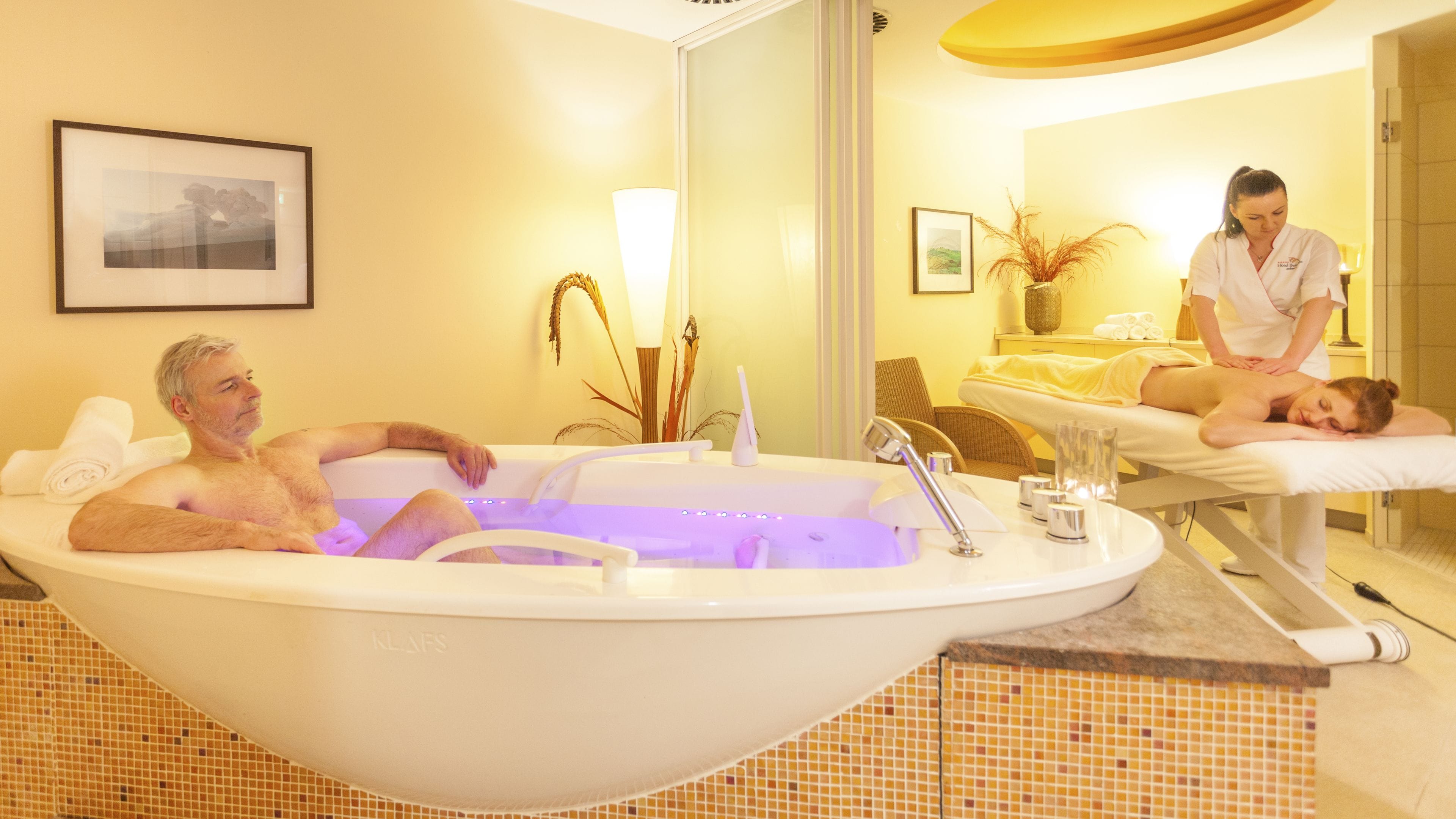 A spa room with a large bathtub, a massage table, and a massage therapist attending to a client.