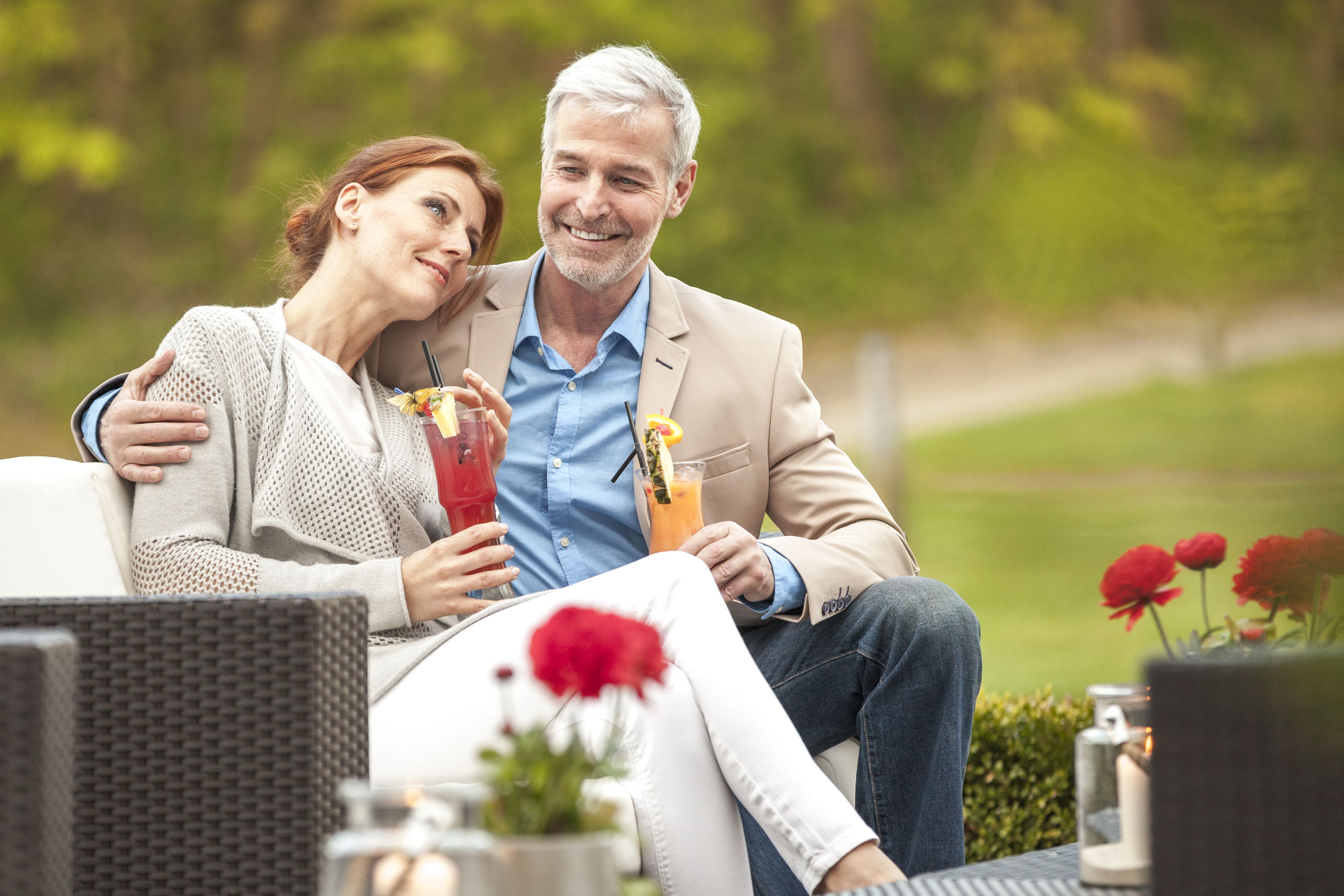 A couple sitting on a wicker chair outdoors with drinks, surrounded by greenery and flowers.