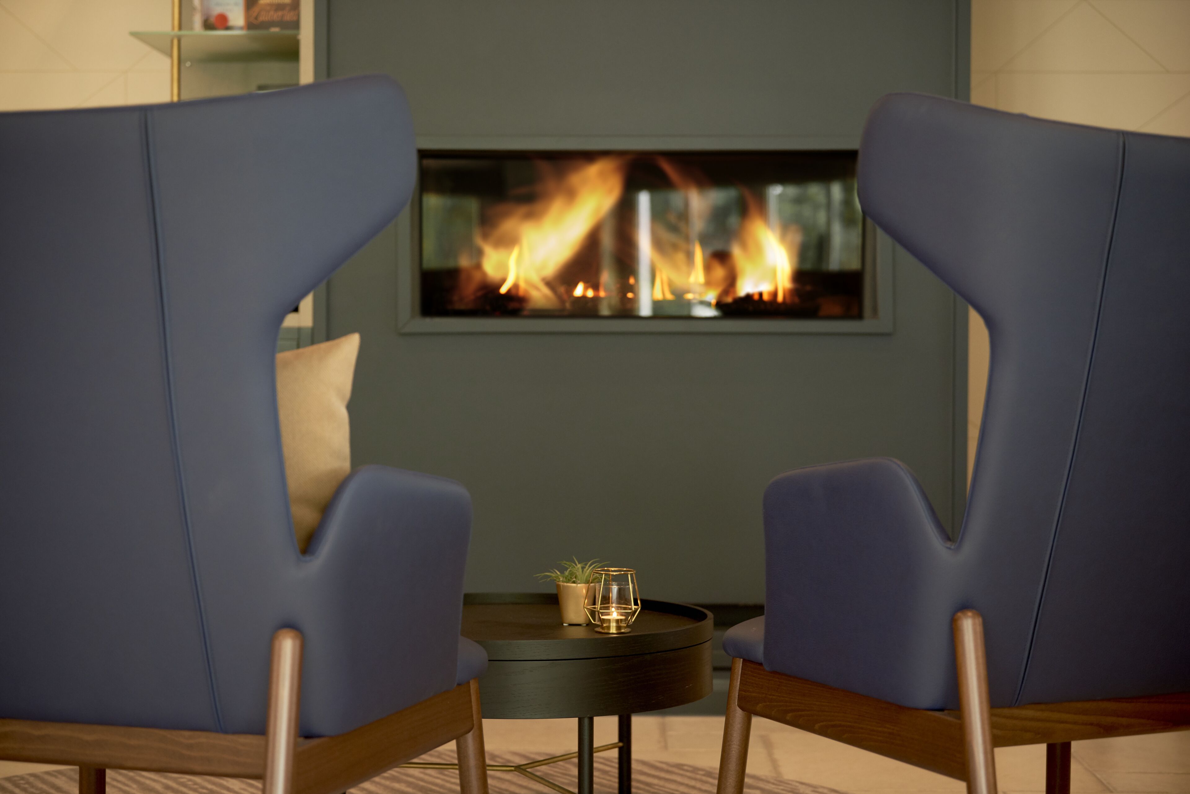 Two blue armchairs facing a fireplace with a potted plant and candle on a small table.