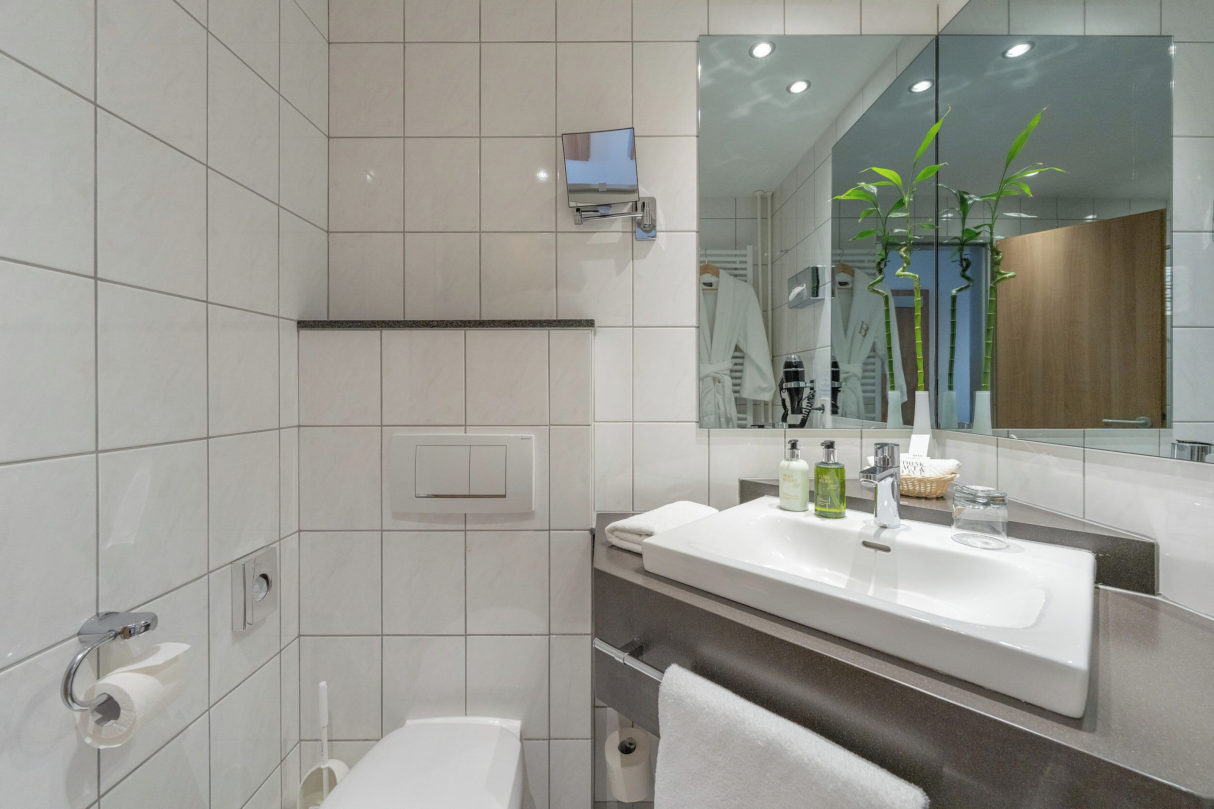 A modern bathroom with a large mirror, white tiles, a sink with toiletries, and a towel rack.