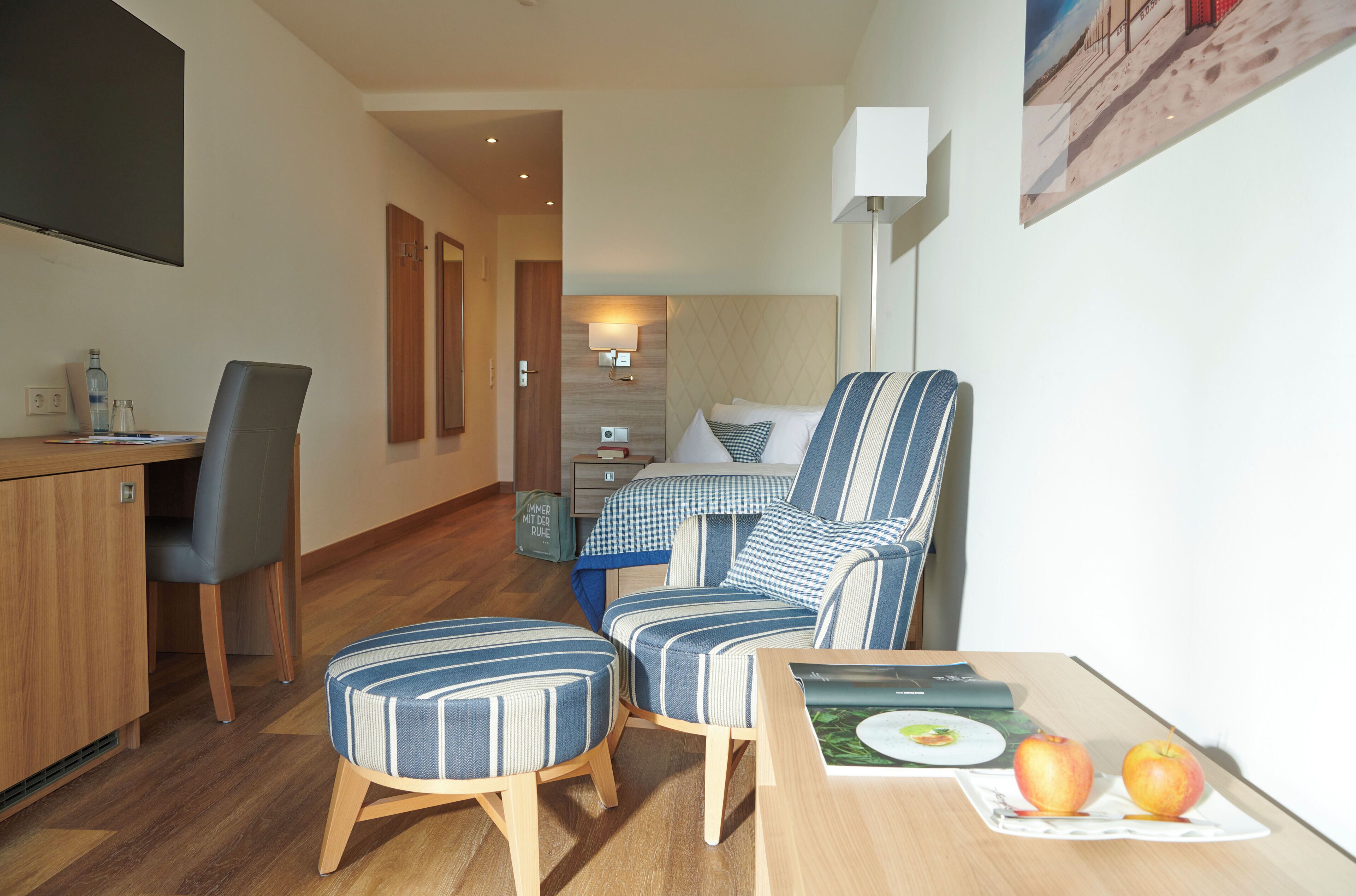 A hotel room with a bed, a desk with a chair, a small table with apples, and a painting on the wall.