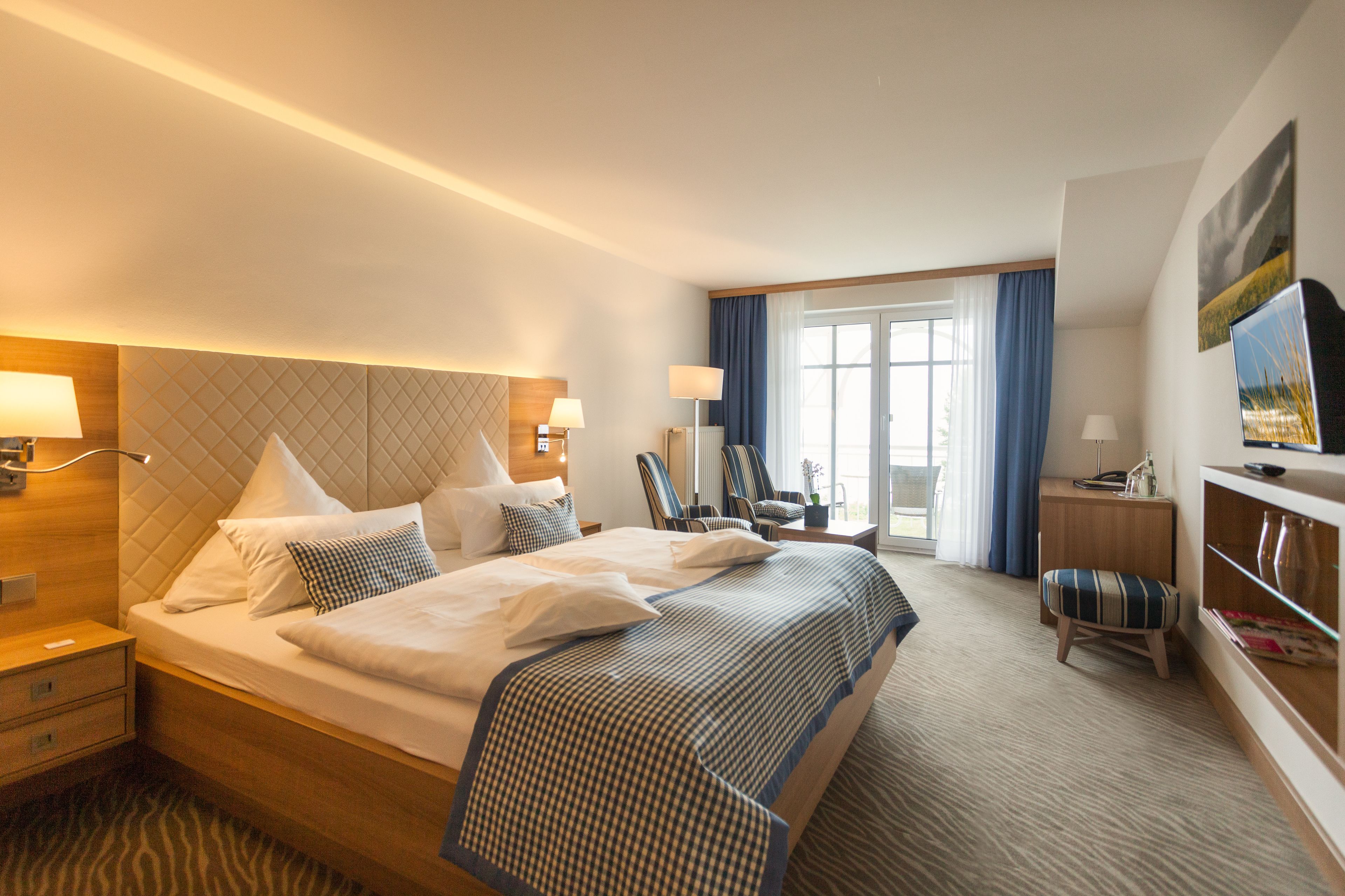 A hotel room with a large bed, bedside tables, a desk, a chair, a television, and a window with curtains.