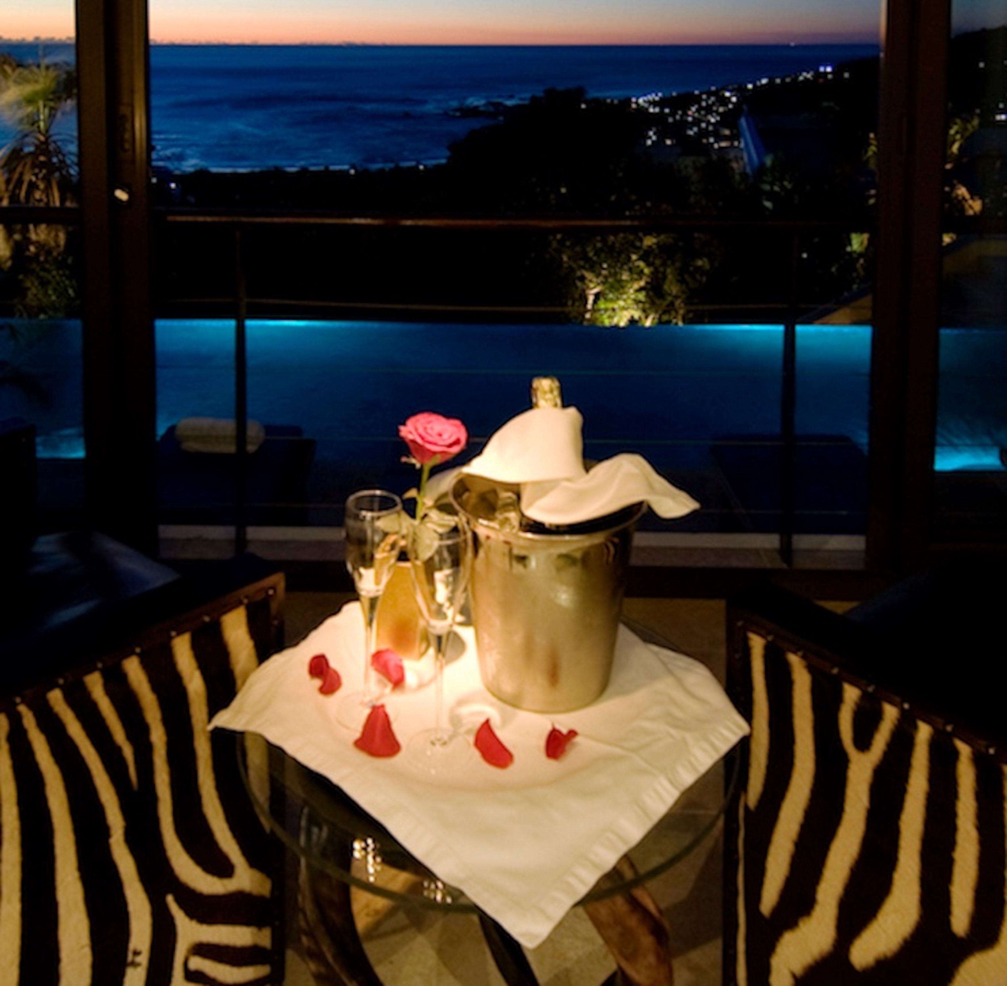 A table with a bottle, glasses, and a rose, set against a view of the ocean and city lights.