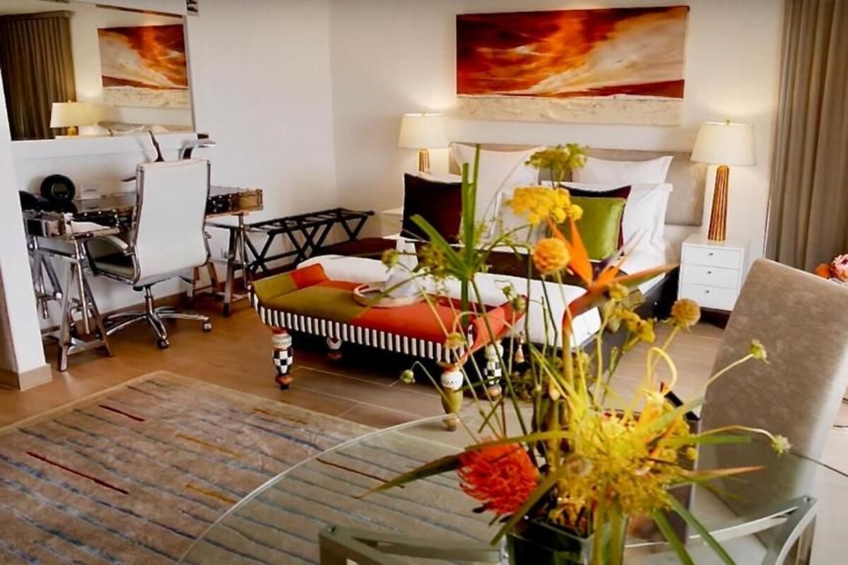 Una habitación de hotel con cama, escritorio, silla, una mesita de vidrio con flores y un sillón rojo.