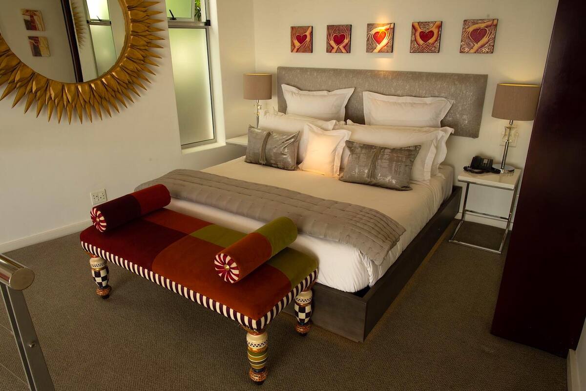 A hotel room with a large bed, a bedside table with a lamp, a round mirror, and heart-themed artwork on the wall.