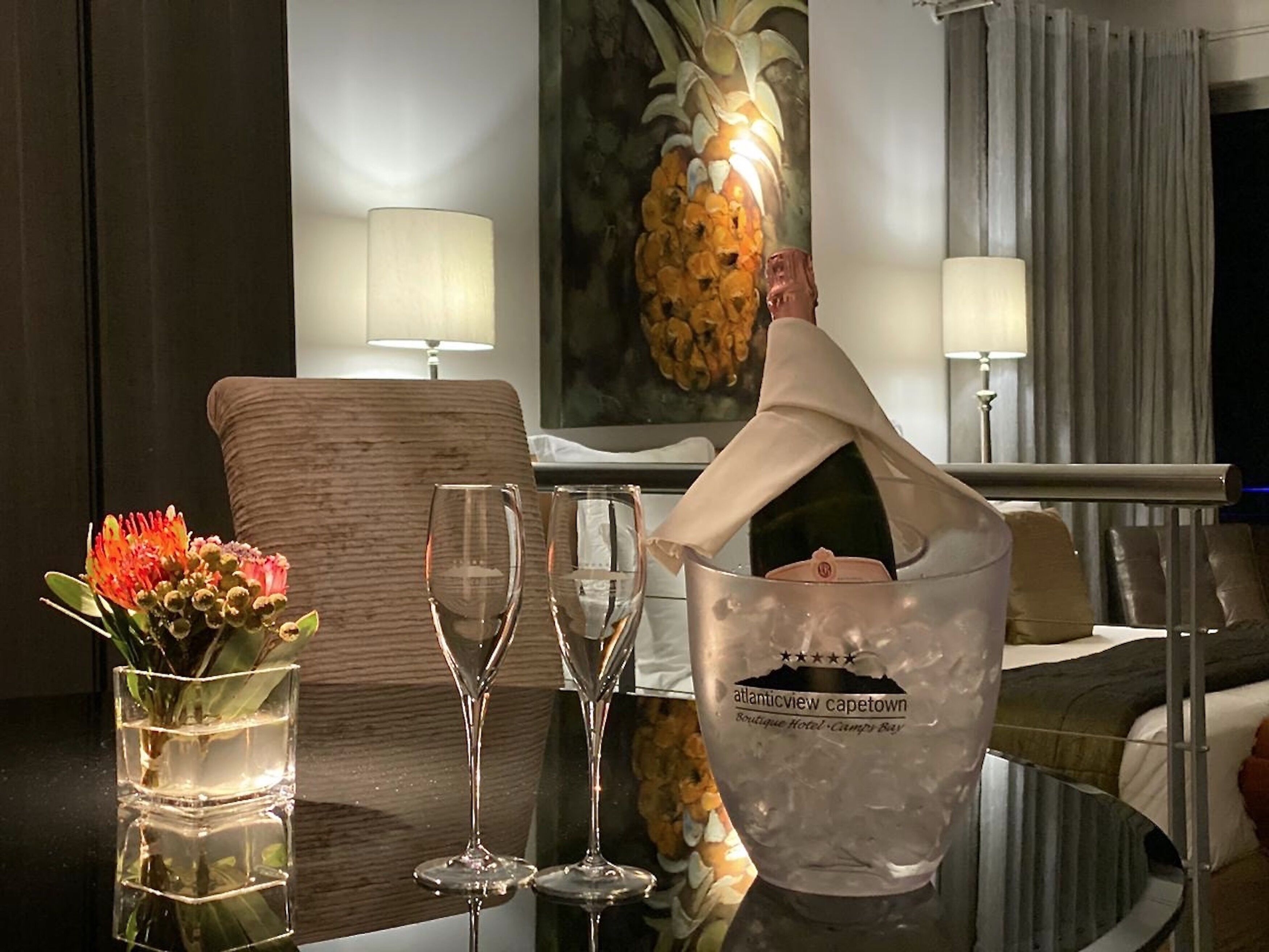A hotel room with a dining table set with wine glasses, a vase with flowers, and a bucket with a bottle of champagne.