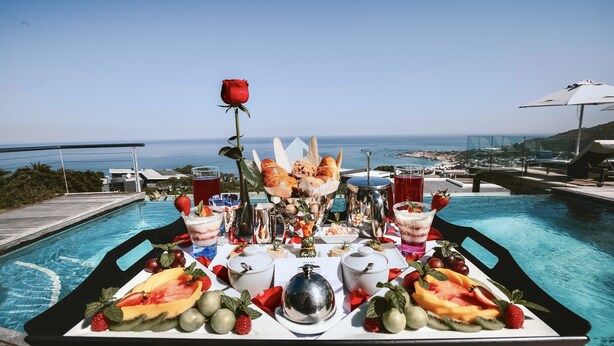 Una mesa con frutas, pasteles y bebidas junto a una piscina con vista al océano.