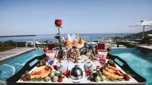 Una mesa con frutas, pasteles y bebidas junto a una piscina con vista al océano.