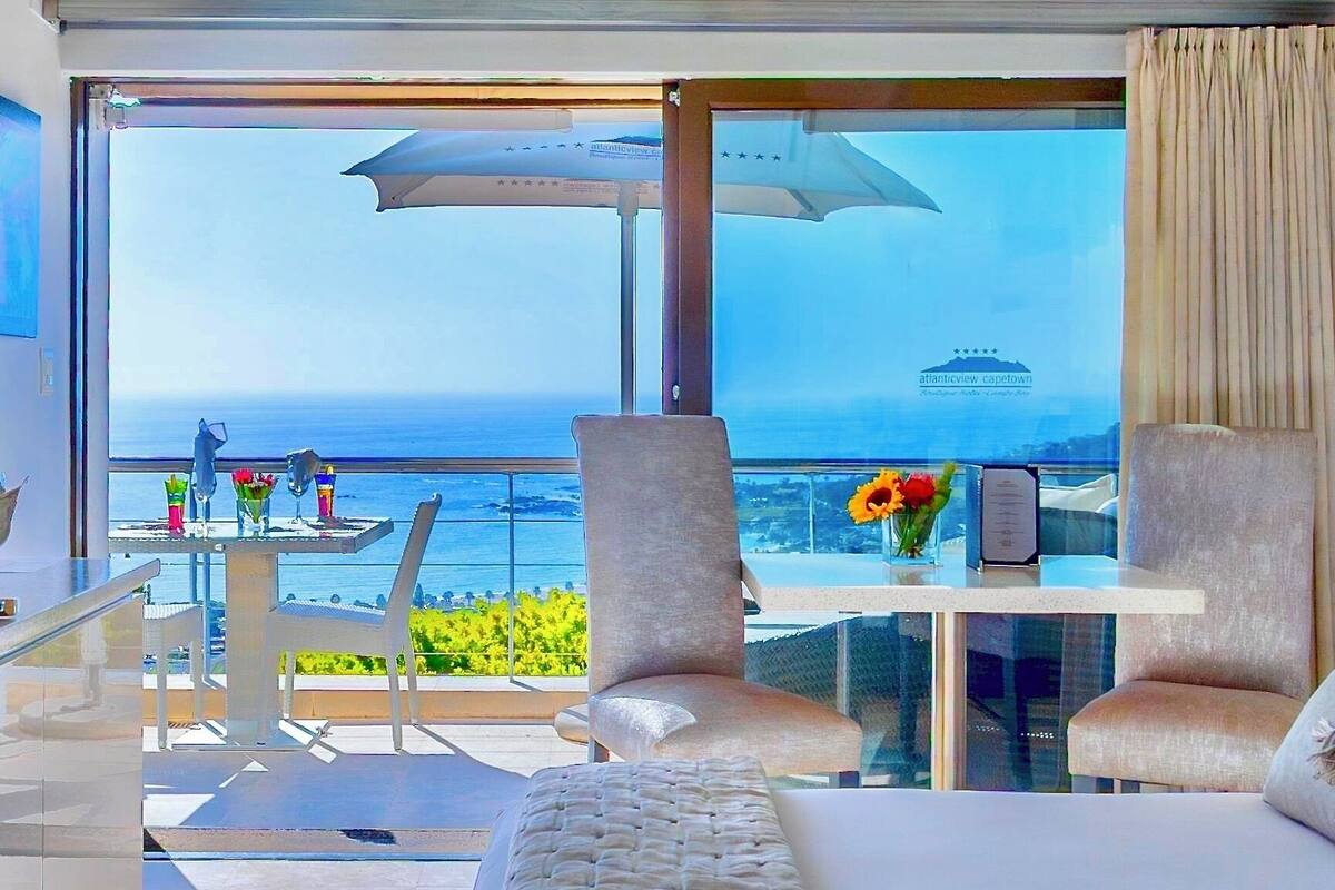 A room with a large window, a table with chairs, a sofa, and a vase with flowers.