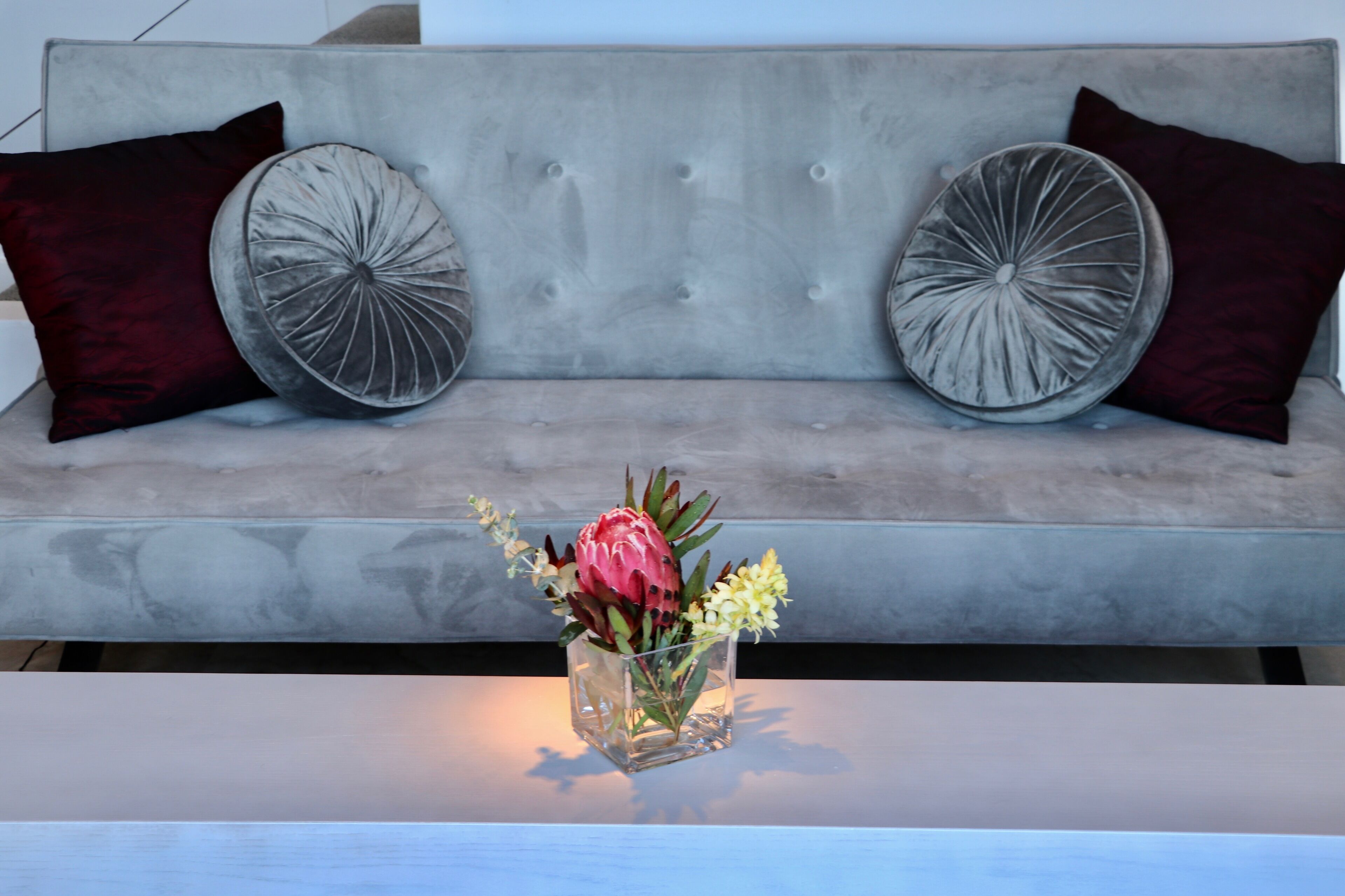 Ein graues Sofa mit zwei runden, grauen Kissen und einer Vase mit Blumen auf einem weißen Tisch im Vordergrund.
