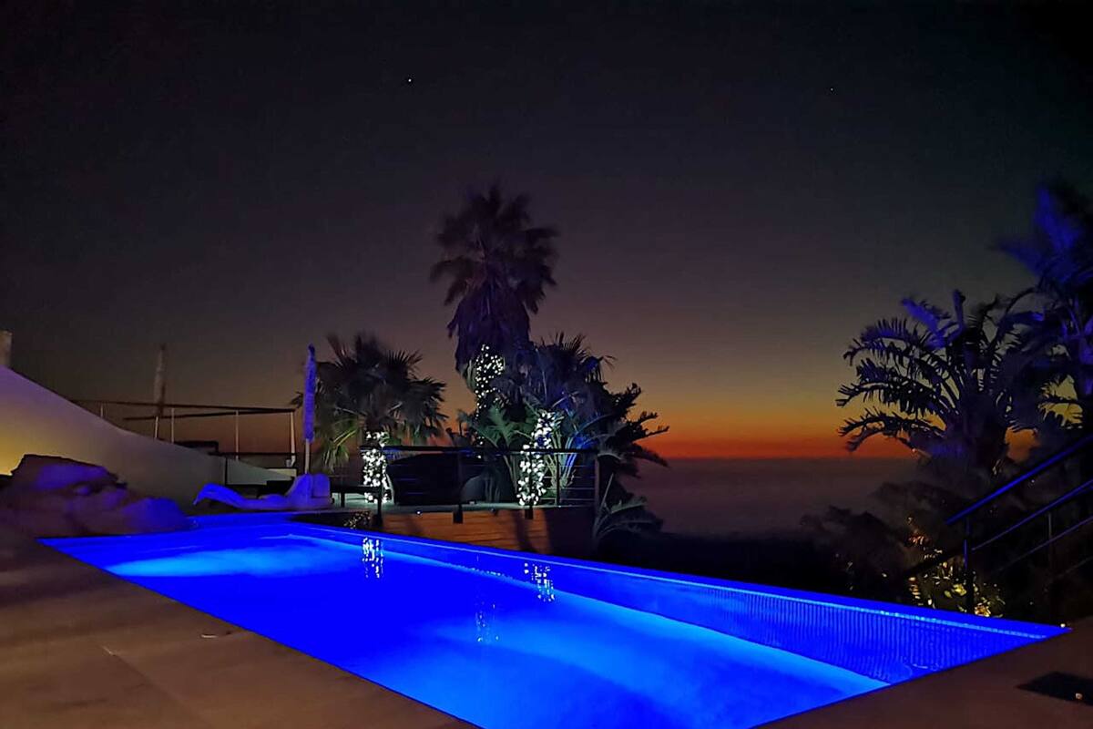 A swimming pool with blue lighting at dusk, surrounded by palm trees and a sunset view.