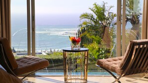 Una habitación moderna con vista al océano, que incluye dos sillones de cuero, una mesa de vidrio con un jarrón de flores y un amplio ventanal con vistas al mar.