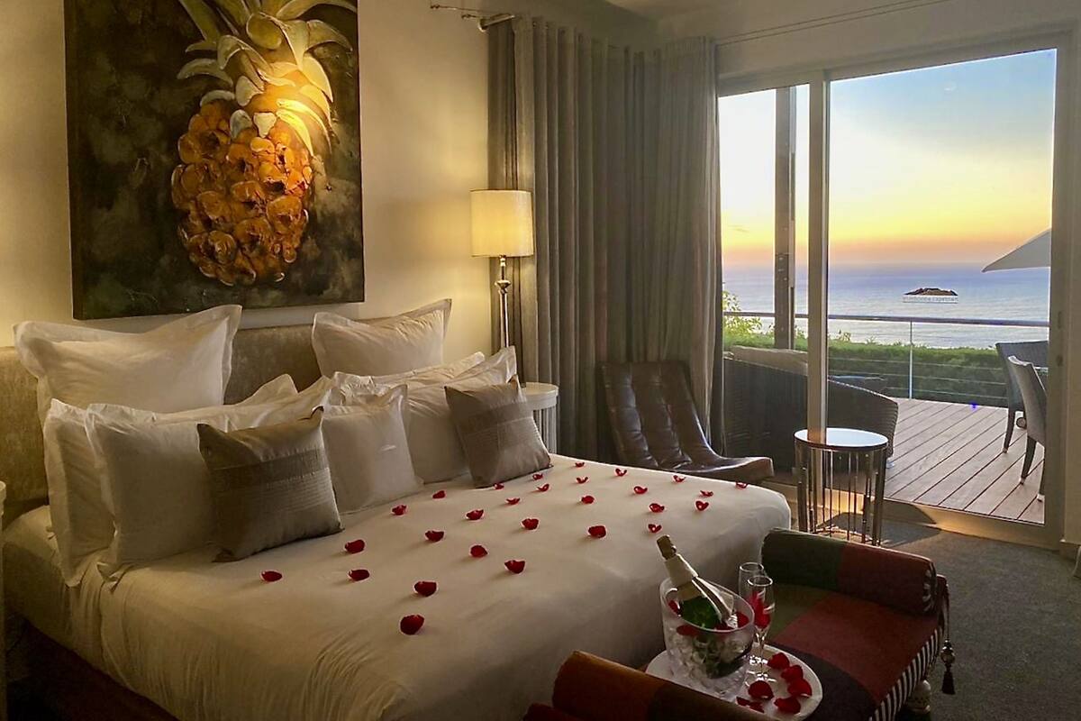 A hotel room with a large bed, a painting of a pineapple, a view of the ocean, and a balcony with outdoor seating.