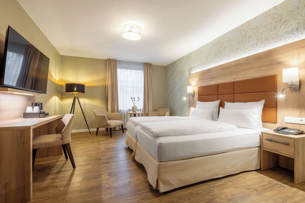 A hotel room with a large bed, a desk with a chair, a TV, a lamp, and a bedside table with a phone.