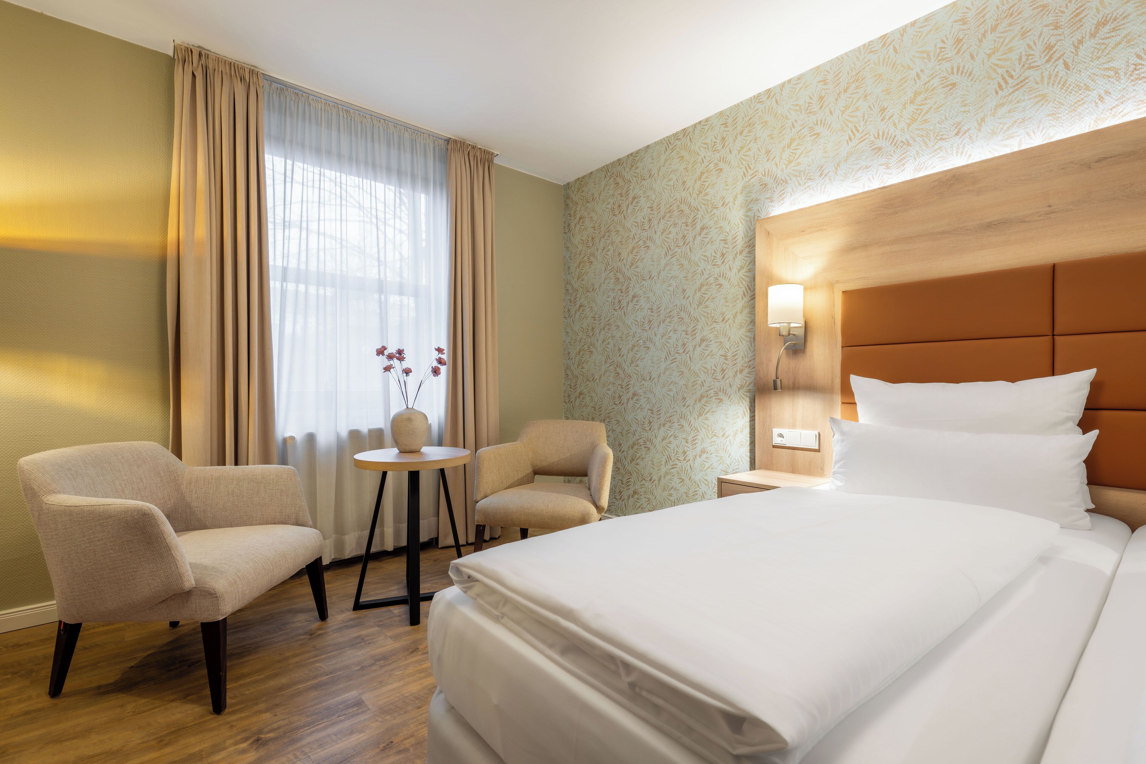 Hotellihuone, jossa on suuri sänky, kaksi nojatuolia, pieni pöytä ja yöpöytä, jossa on lamppu.