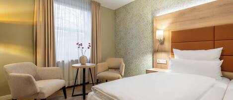 Habitación de hotel con una cama grande, dos sillones, una mesita y una mesita de noche con una lámpara.
