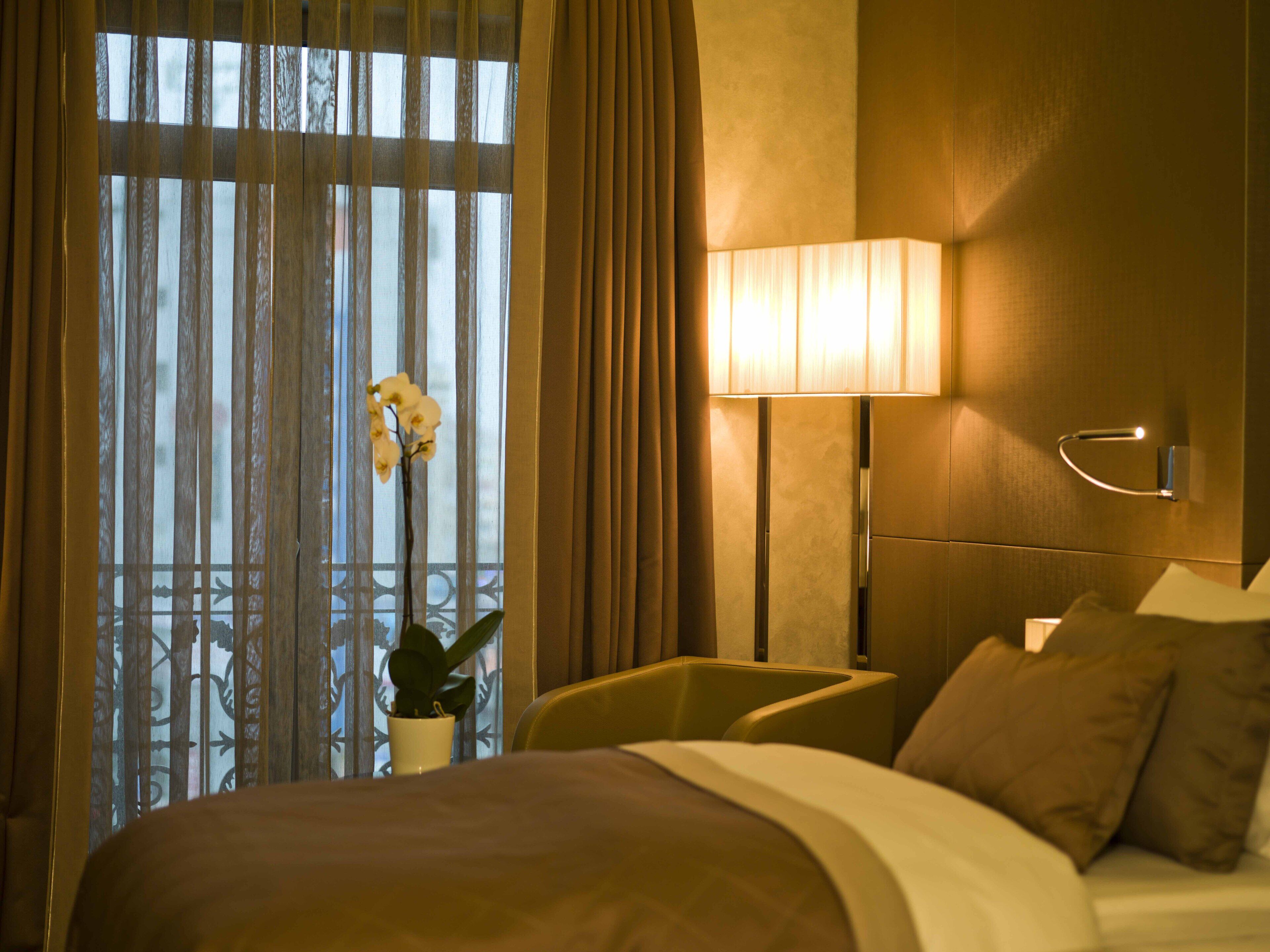 A hotel room with a bed, a bedside lamp, a potted plant, and a window with curtains.