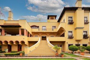 Un edificio de varios pisos con una escalera central que conduce a la entrada, rodeado de follaje y plantas en macetas.