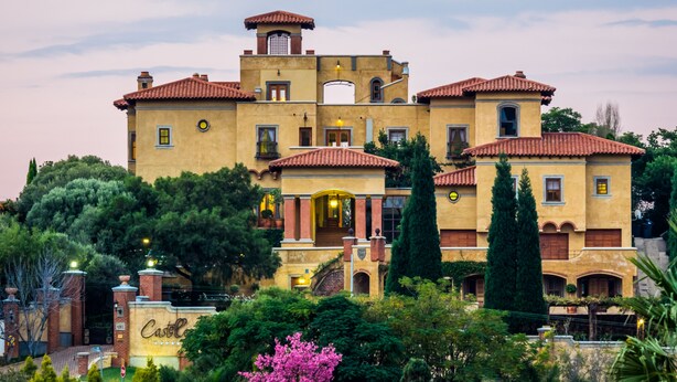 A large, multi-story building with a yellow facade and red-tiled roofs, surrounded by lush greenery and a clear sky. - Castello di Monte (Pretoria)