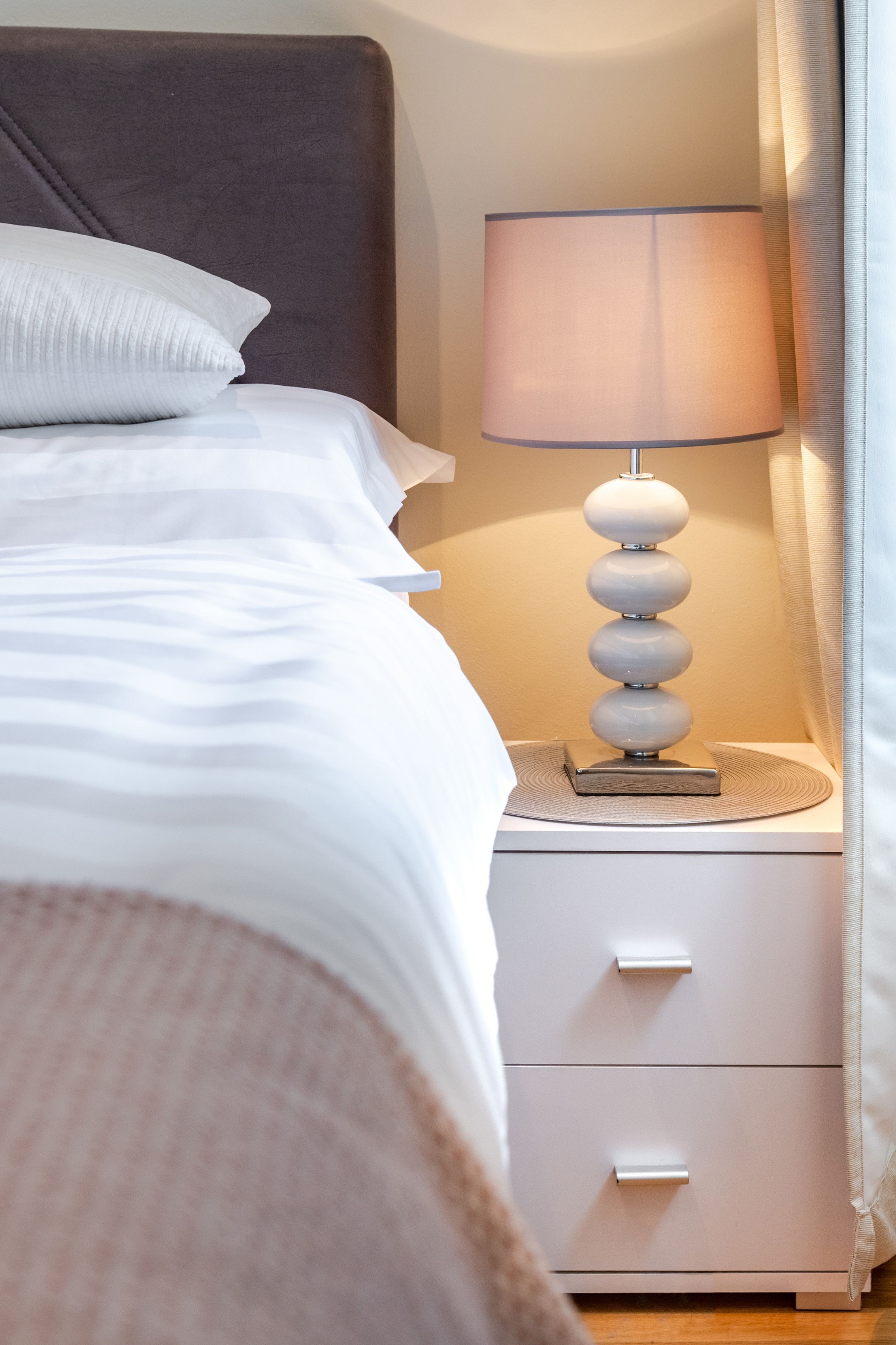 A neatly made bed with white bedding, a white bedside table with a lamp, and a white drawer.