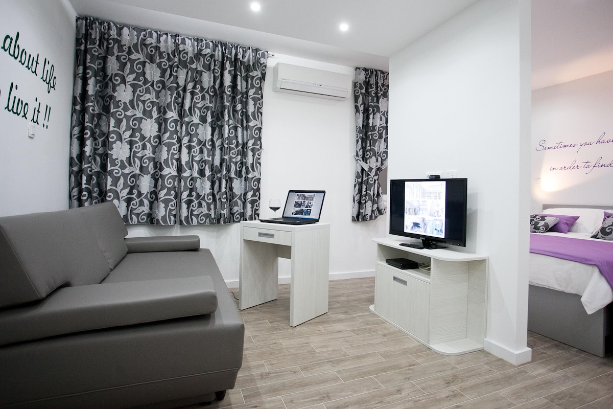 A modern hotel room with a grey sofa, a desk with a laptop and a flat-screen TV, a bed with a purple blanket, and decorative wall art.