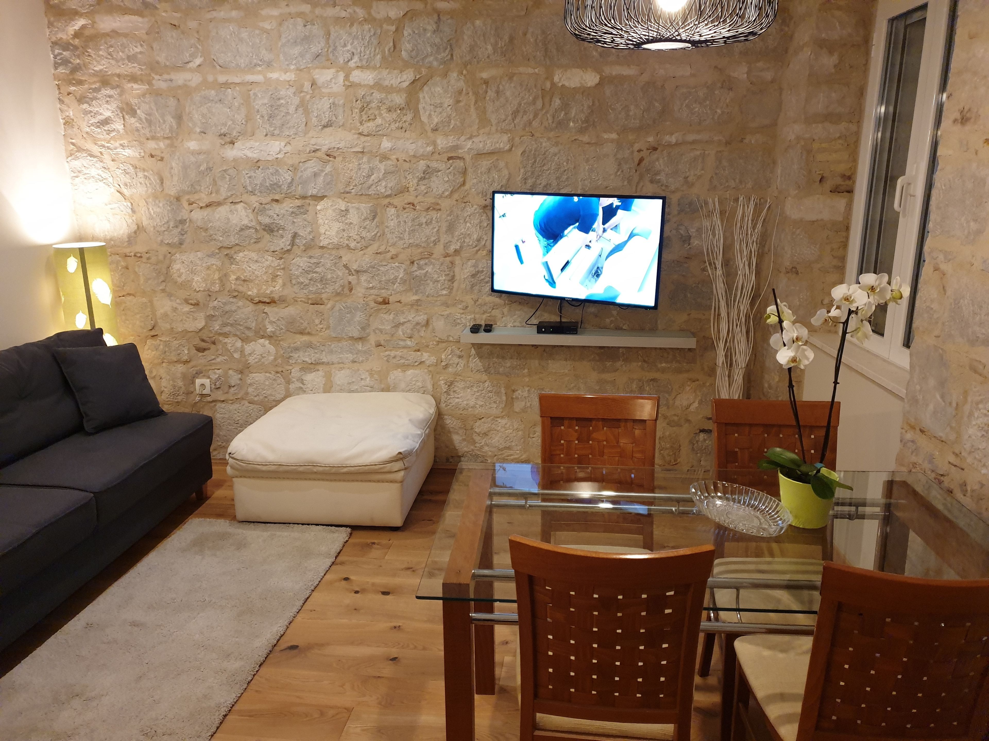 Una habitación con paredes de piedra, un sofá, un reposapiés, una mesa de comedor, sillas, un televisor y una lámpara colgante.