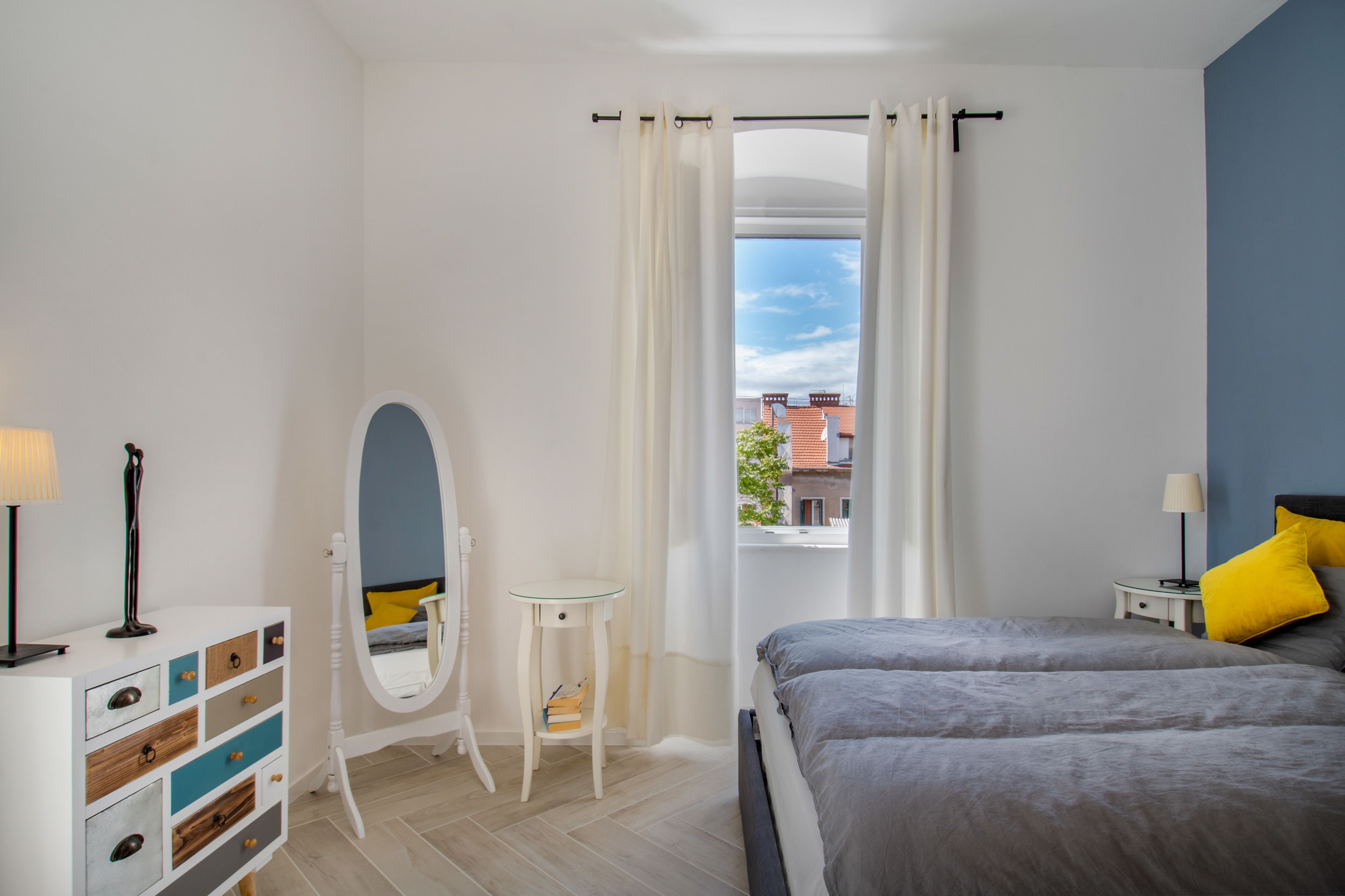 A bedroom with a bed, a dresser, a mirror, a small table, and a window with curtains.