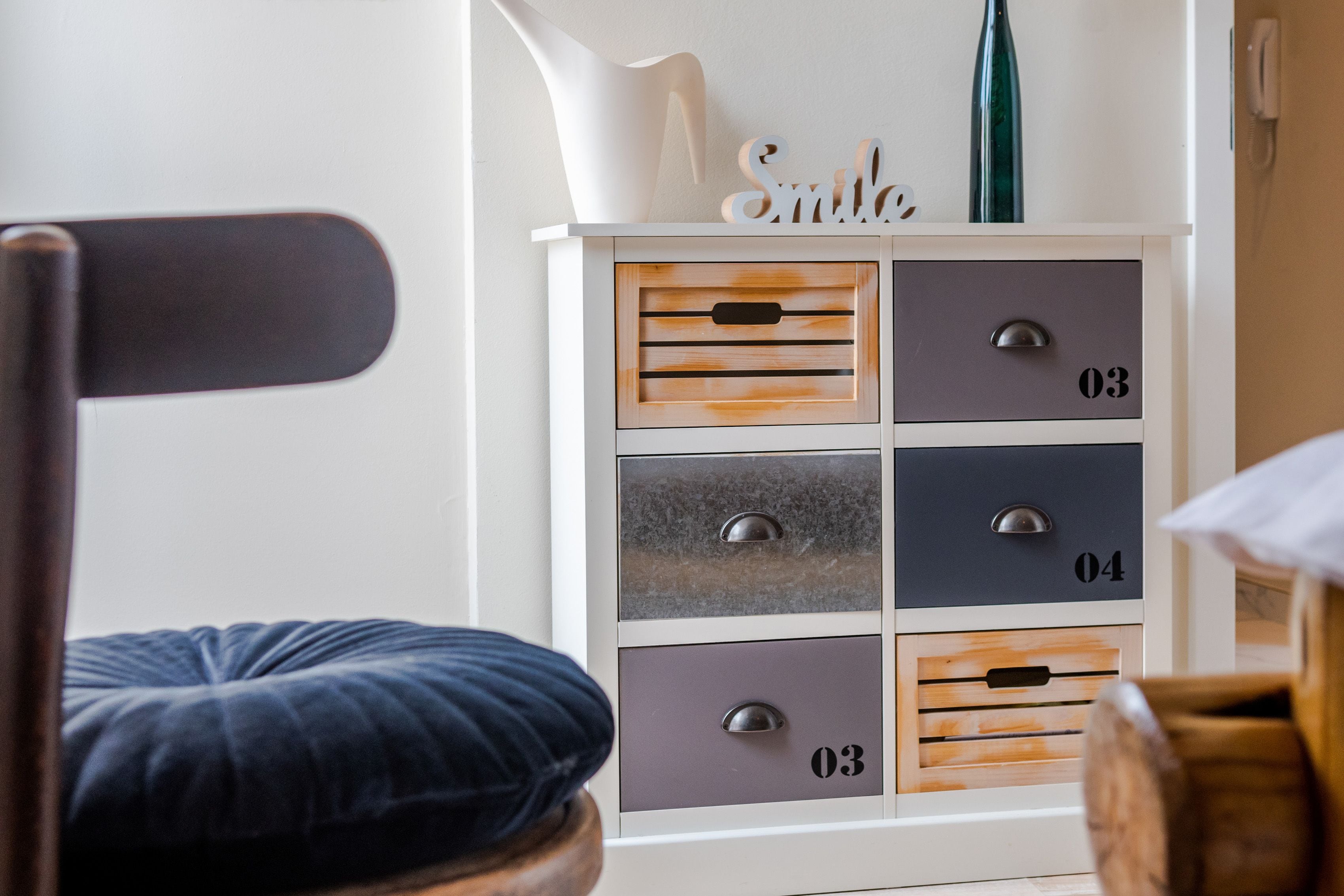 Una habitación con un armario blanco con cajones etiquetados como 03 y 04, un cojín azul y una silla de madera.