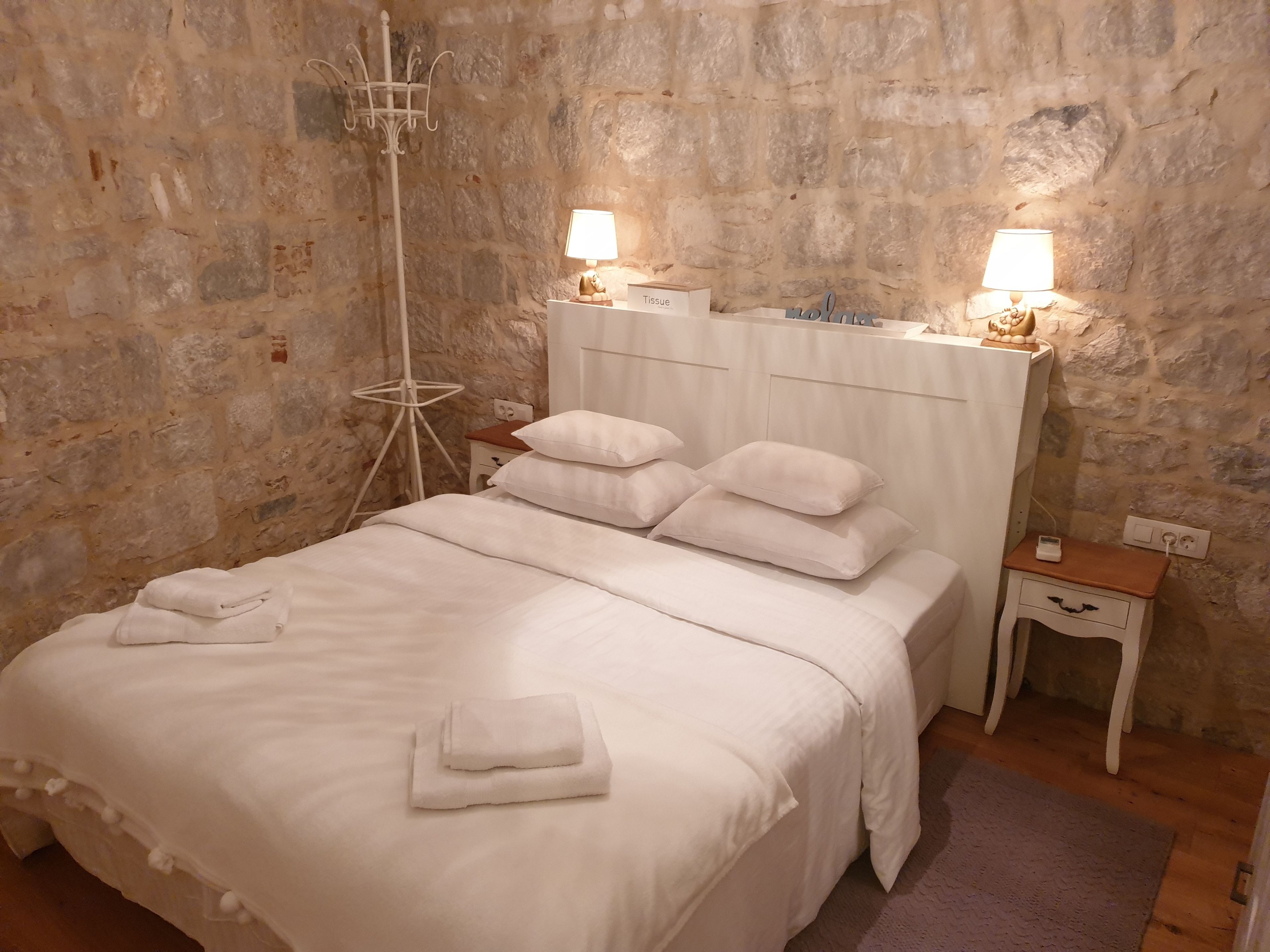 Un lit bien fait, avec du linge de lit blanc et des oreillers, une table de chevet avec une lampe, et un mur à la texture de pierre.