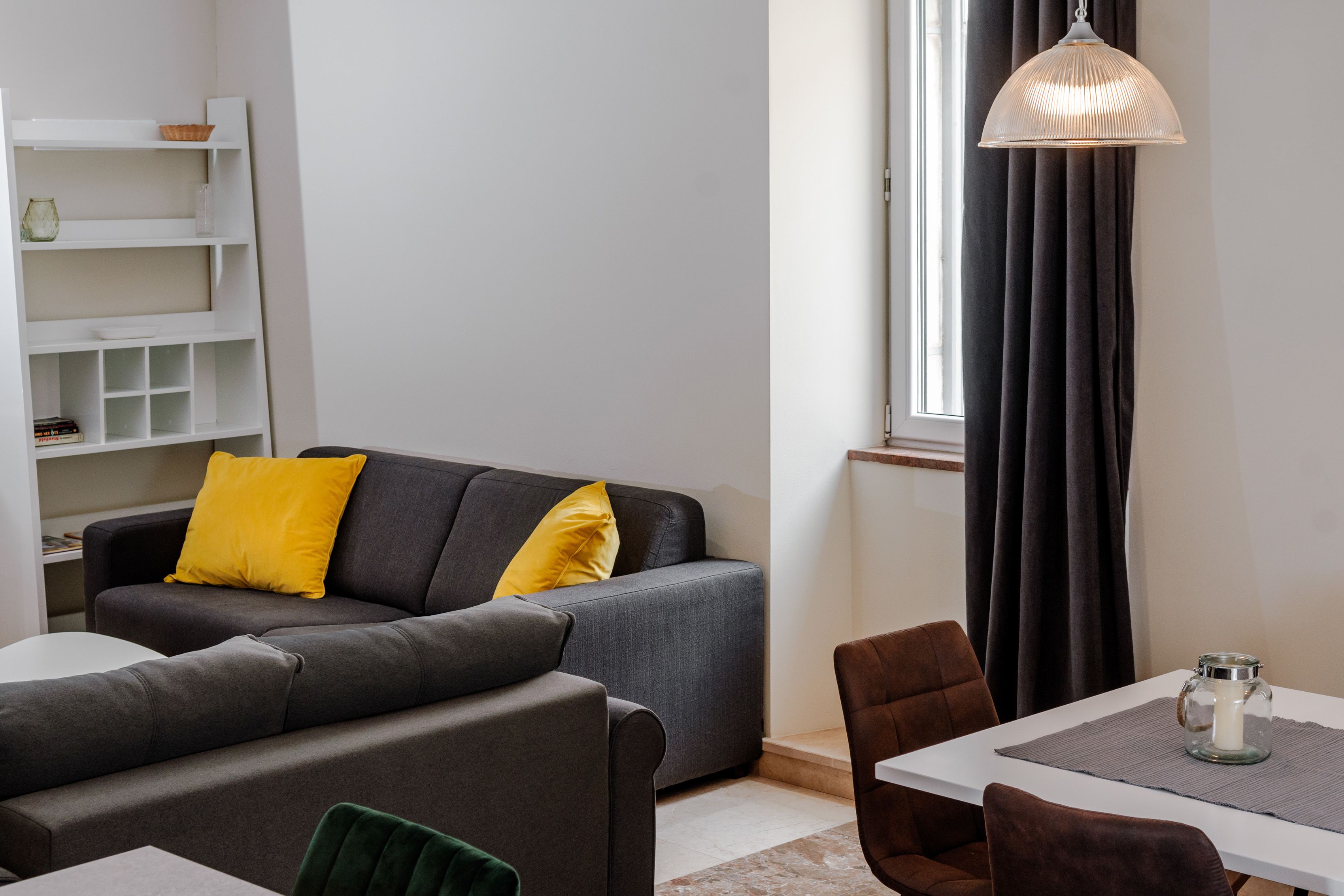 A modern living room with a grey sofa, yellow cushions, a dining table with chairs, and a hanging light fixture.